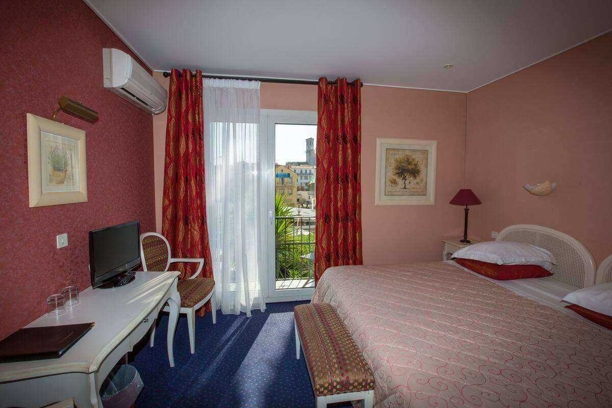 A hotel room with a bed, a desk with a computer, a chair, a window with curtains, and a balcony view.