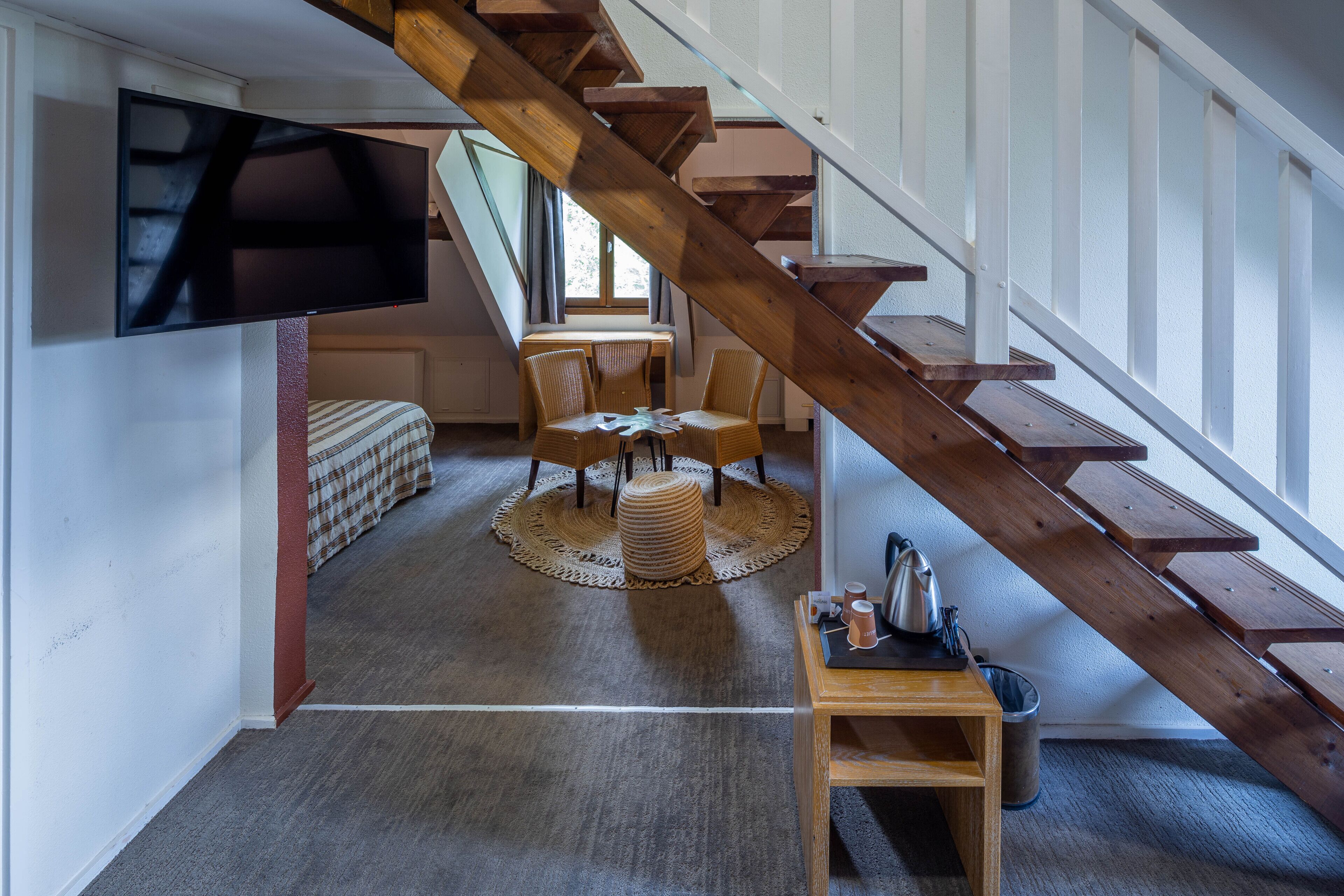 Un intérieur moderne avec un escalier en bois, un téléviseur à écran plat fixé au mur, un coin repas avec une table ronde et des chaises, et un coin salon confortable agrémenté d’un tapis à motifs.