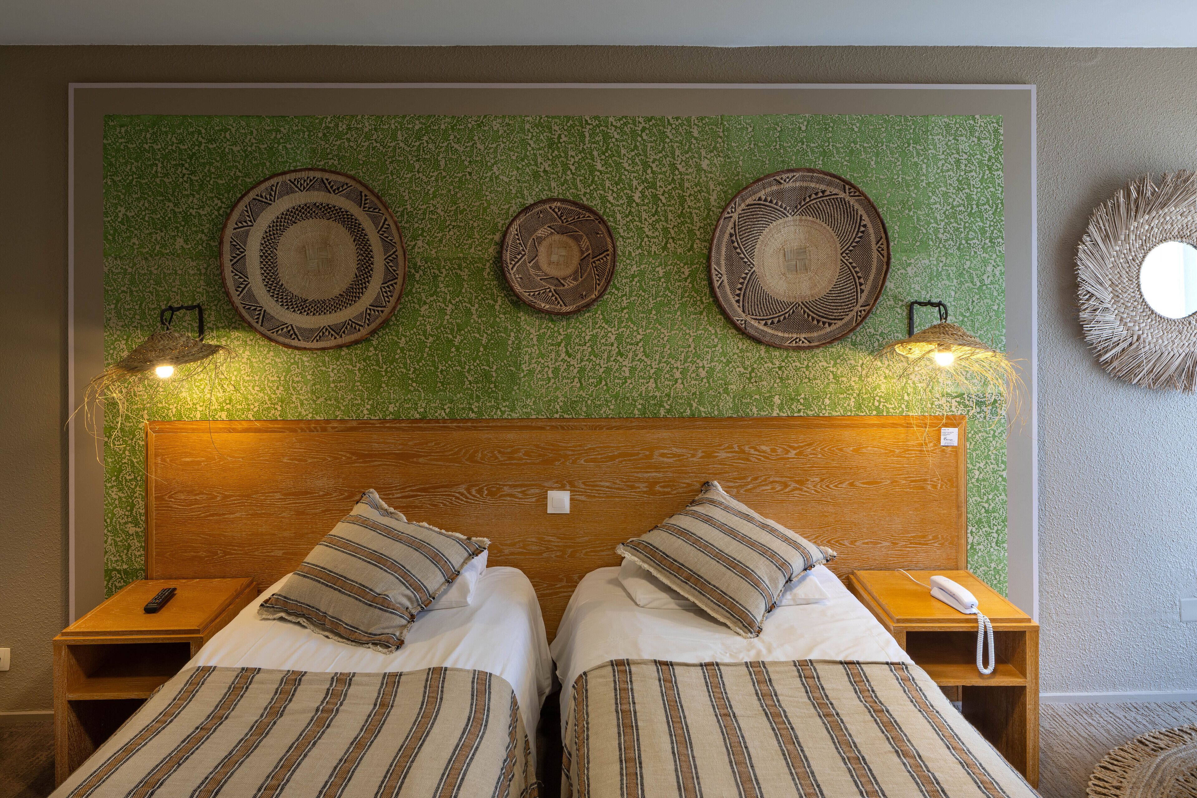A hotel room with two beds, a wooden headboard, and a green textured wall. There are decorative plates above the beds and a woven wall hanging on the right.