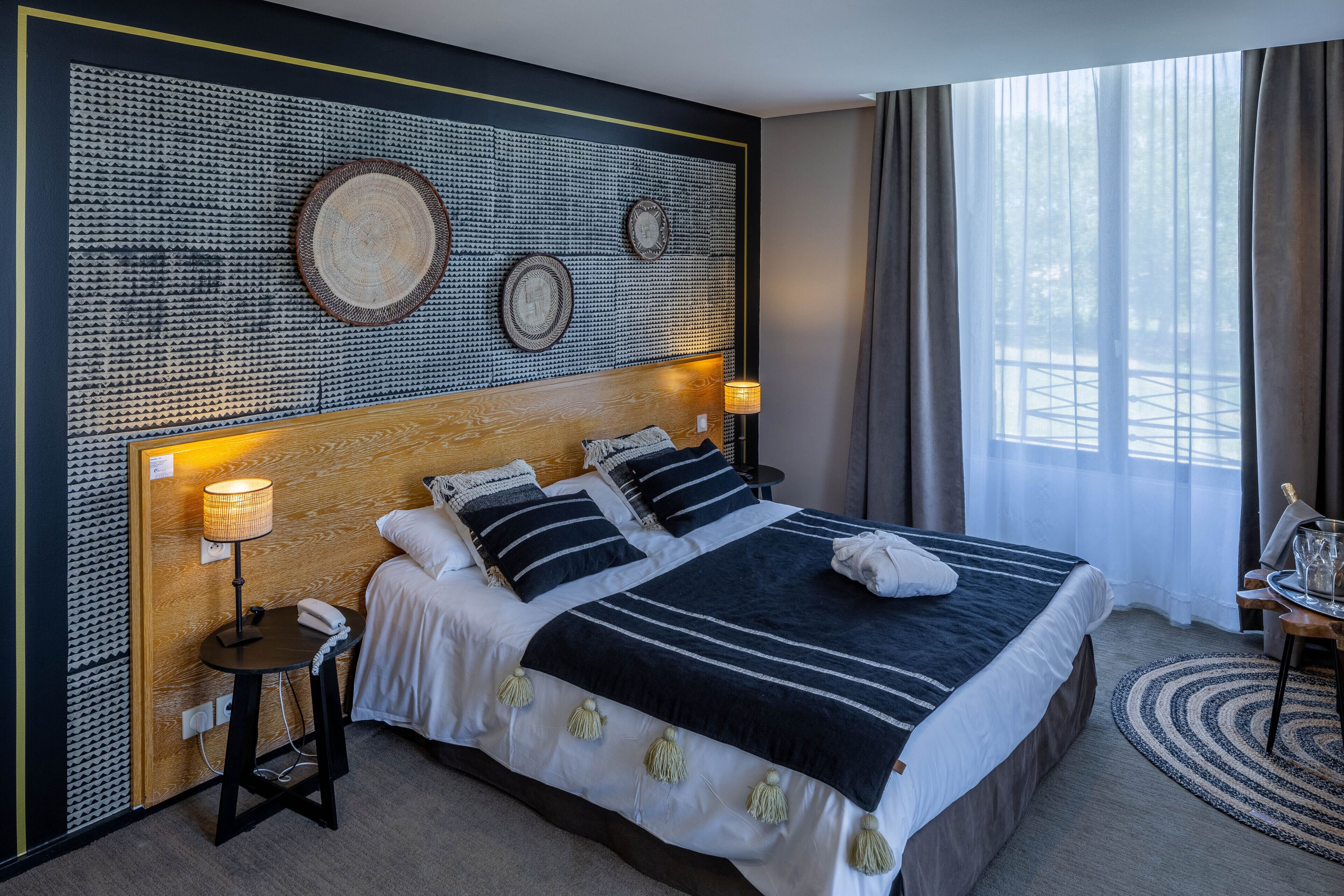 Ett hotellrum med en stor säng, en sänggavel dekorerad med konst, en sänglampa och utsikt över omgivningarna genom ett fönster.