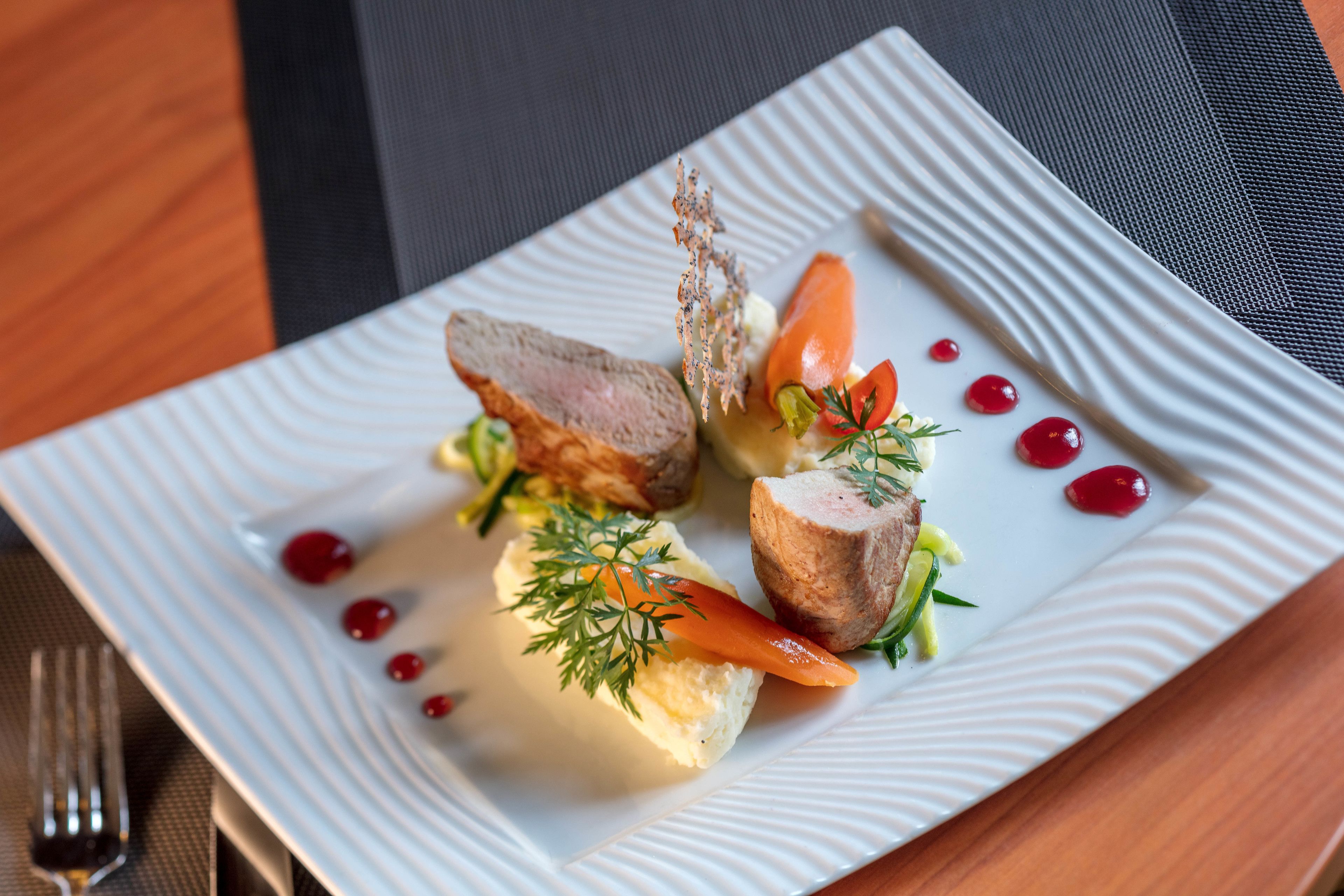 A white plate with a piece of meat, a carrot, and some greens on a textured tablecloth.