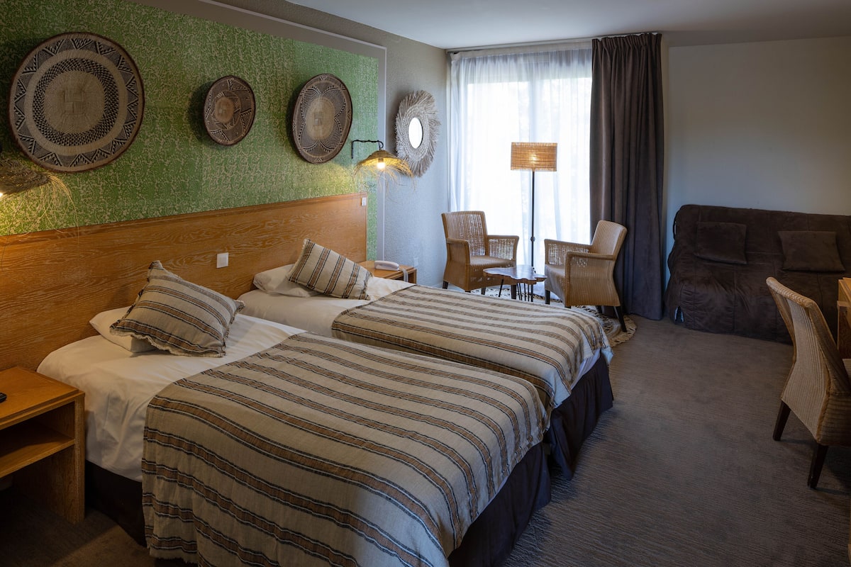 Une chambre d’hôtel avec deux lits, une tête de lit en bois, un mur vert à texture et une grande fenêtre avec des rideaux.