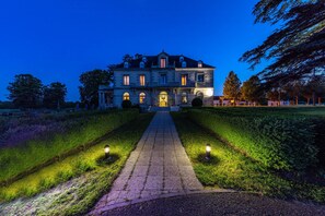 Een groot, historisch gebouw met een symmetrische gevel, een centrale ingang en een verzorgde tuin met een geplaveid pad dat naar de ingang leidt.