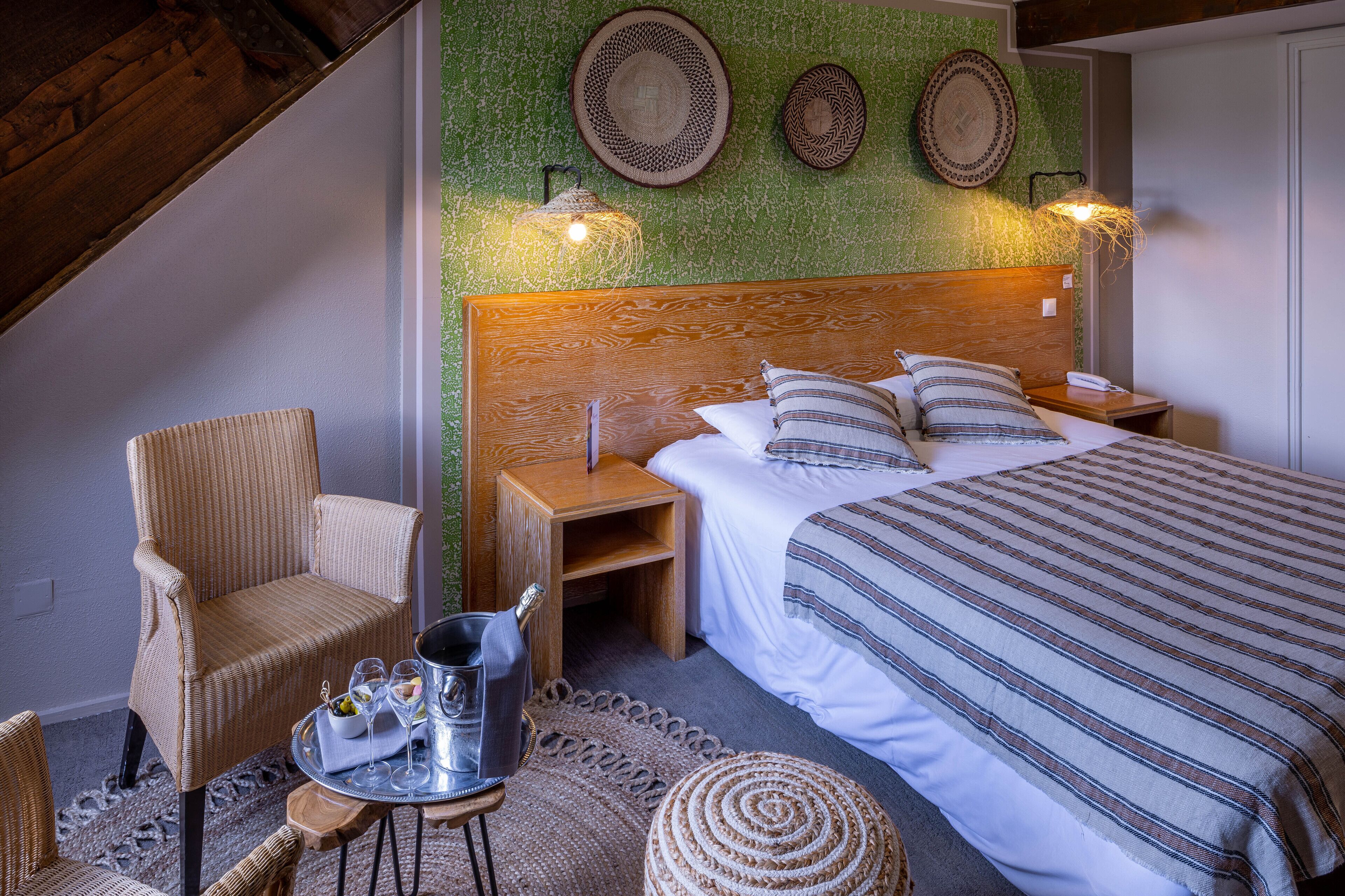 Ein Schlafzimmer mit einem Holz-Kopfteil, einem Bett mit gestreifter Bettwäsche, zwei Nachttischen, einem Korbstuhl, einem kleinen runden Tisch mit einem Tablett und Gläsern sowie einer Flasche und Wandleuchten.