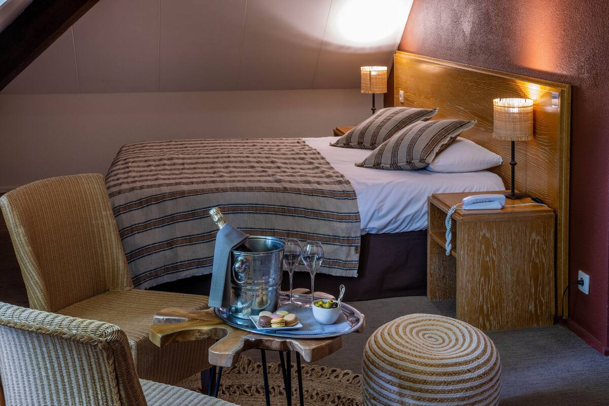 Une chambre d’hôtel confortable avec un grand lit, un fauteuil en osier, une petite table sur laquelle se trouve un plateau de collations et une bouteille de champagne.