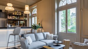 A modern living room with a grey sectional sofa, a bar area with shelves of bottles, and large windows with a view of greenery. - Garrigae Manoir de Beauvoir (Mignaloux-Beauvoir)