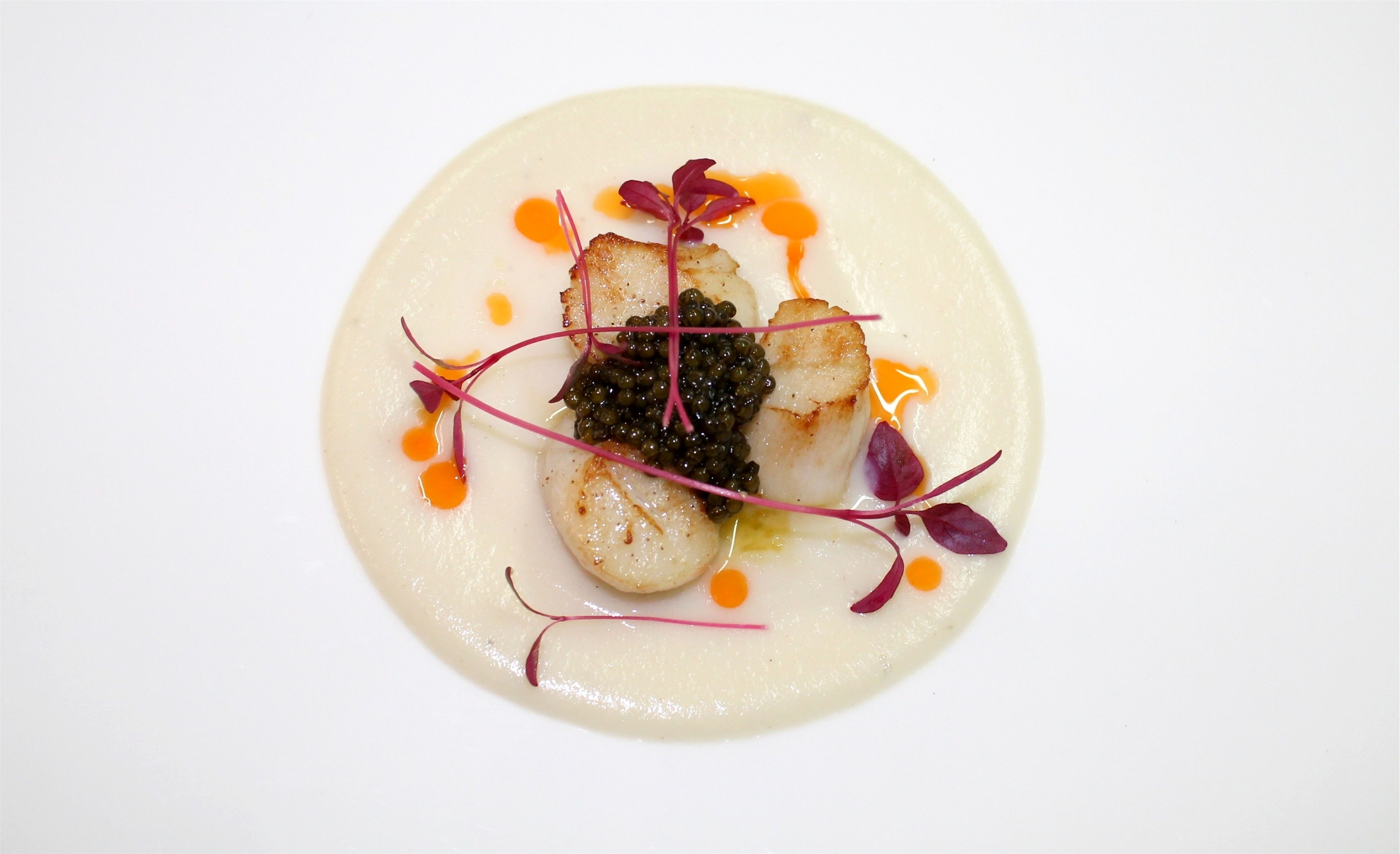 Une assiette de mets comprenant des Saint-Jacques poêlées, du caviar et diverses garnitures.