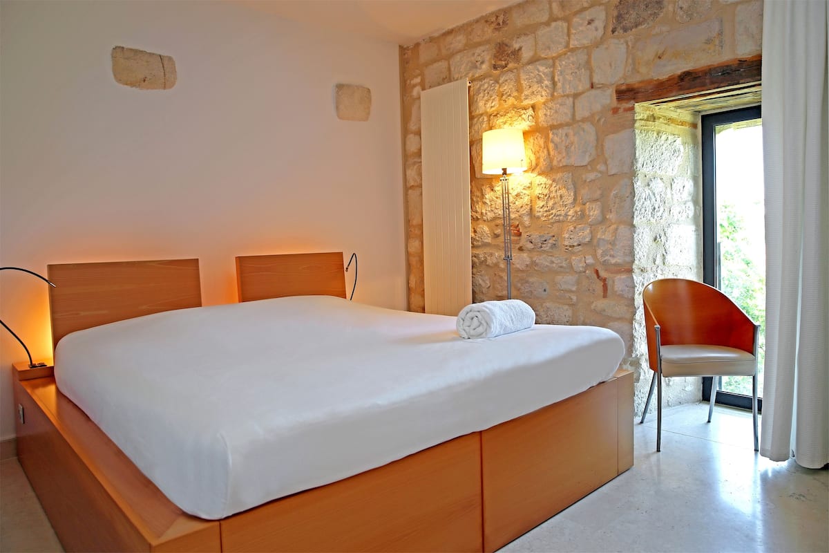 A bedroom with a large bed, a stone wall, a chair, and a lamp.