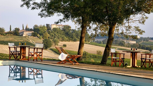 Zona junto a la piscina con sillas y mesas de madera, vistas despejadas a viñedos y un edificio al fondo.