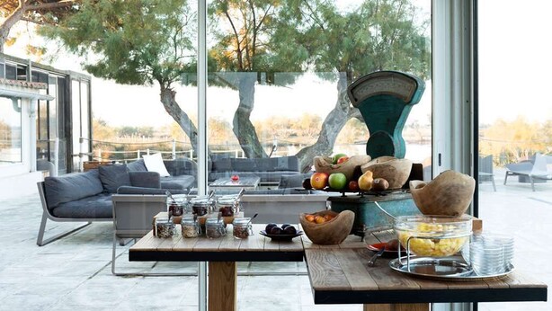 A modern outdoor dining area with a wooden table, seating, and a view of trees and a building in the background. - L'estelle en Camargue (Saintes-Maries-de-la-Mer)