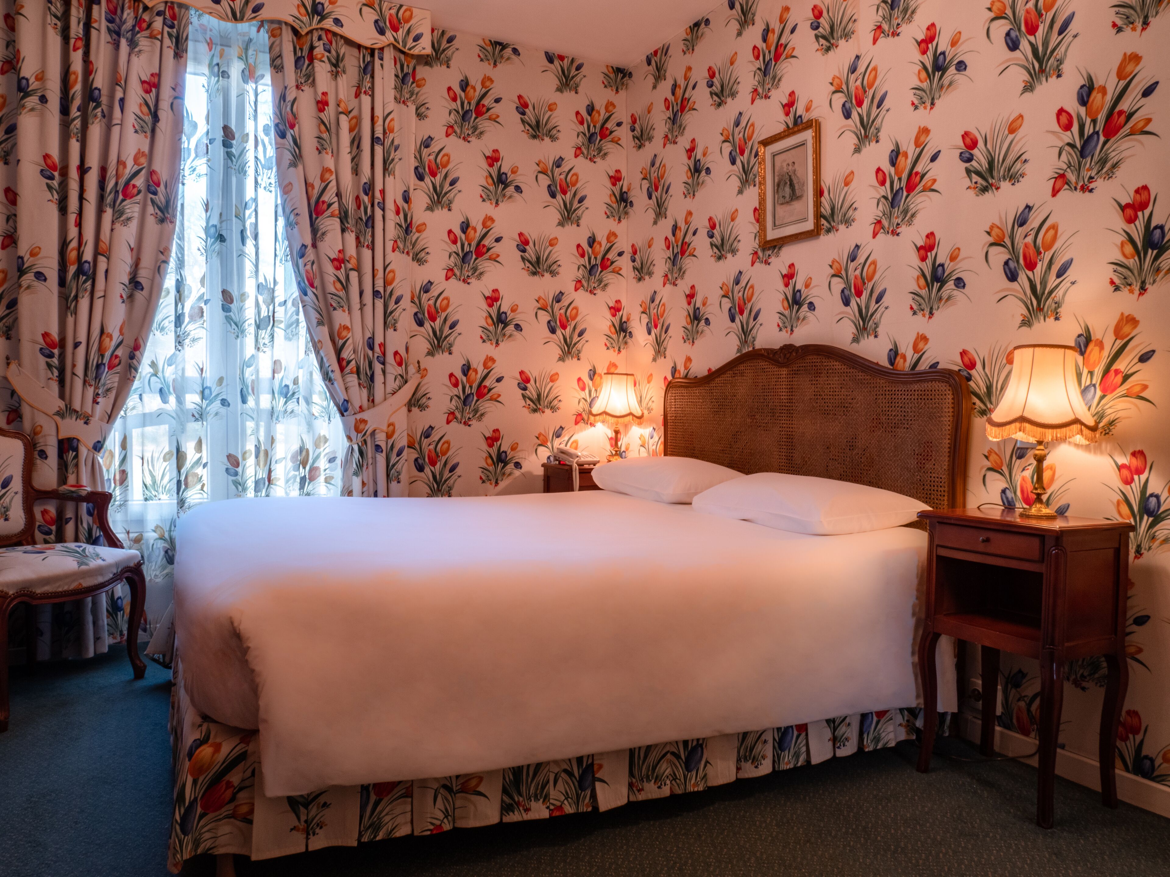 A neatly made bed in a room with floral wallpaper, a wooden headboard, a bedside table with a lamp, and a chair with floral upholstery.