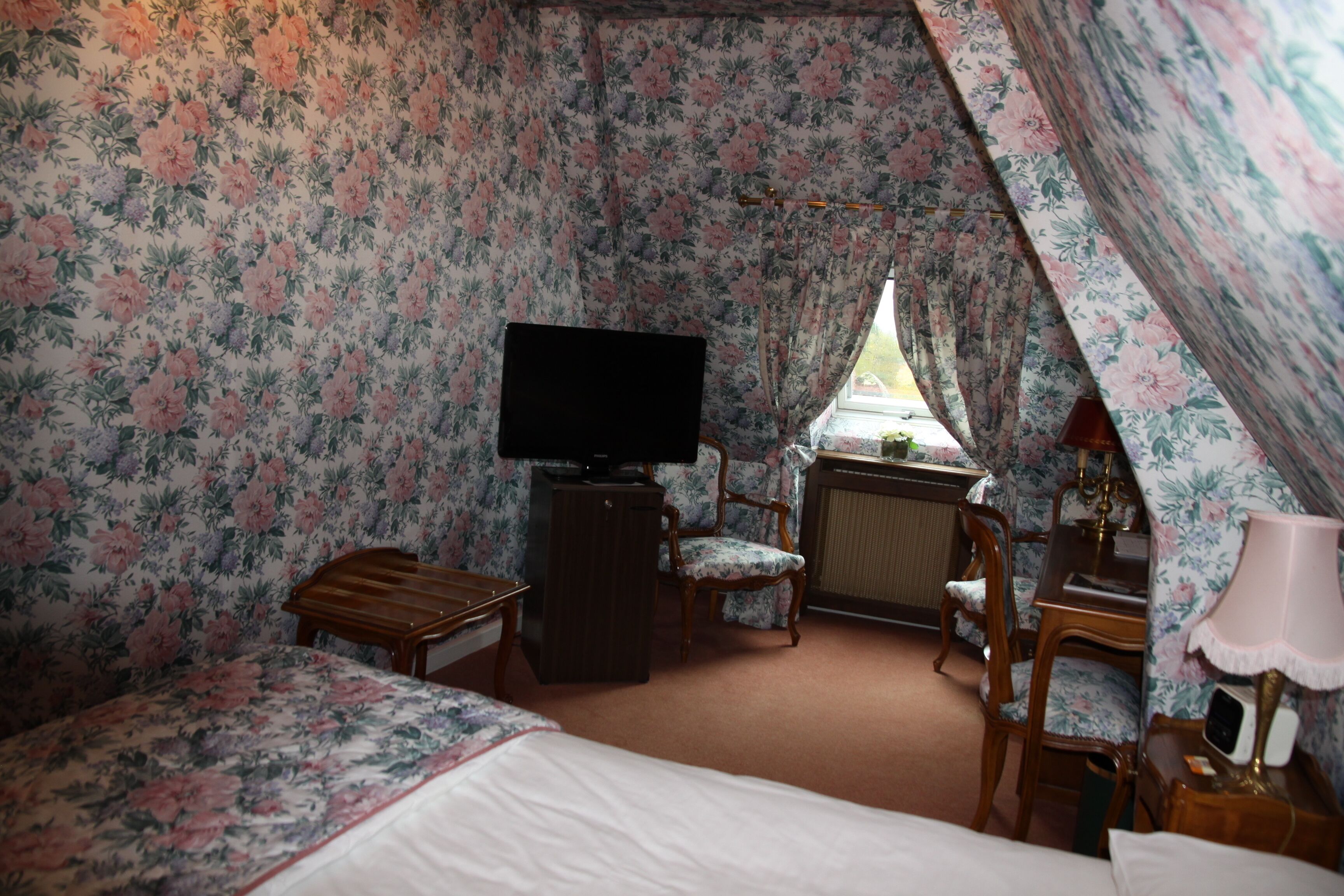 A room with floral wallpaper, a bed, a desk with a lamp, a chair, and a television.