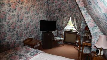 A room with floral wallpaper, a bed, a desk with a lamp, a chair, and a television.