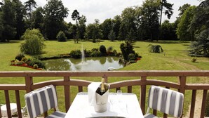 A terrace with a table, two chairs, a bottle of champagne, and a view of a pond surrounded by trees. - Château Des Bondons EI (La Ferte-sous-Jouarre)