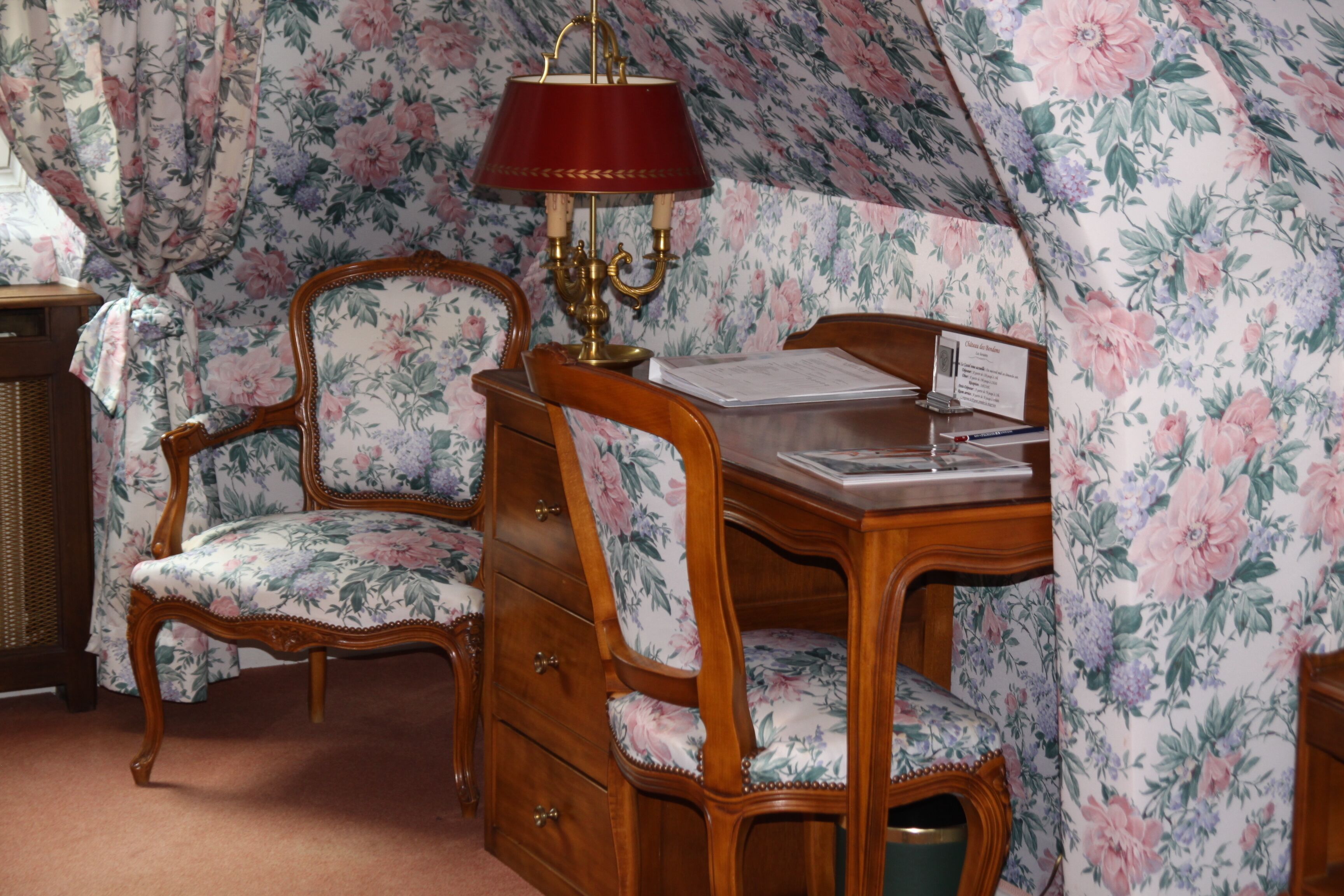 A room with a wooden desk, two chairs, a lamp, and floral wallpaper.