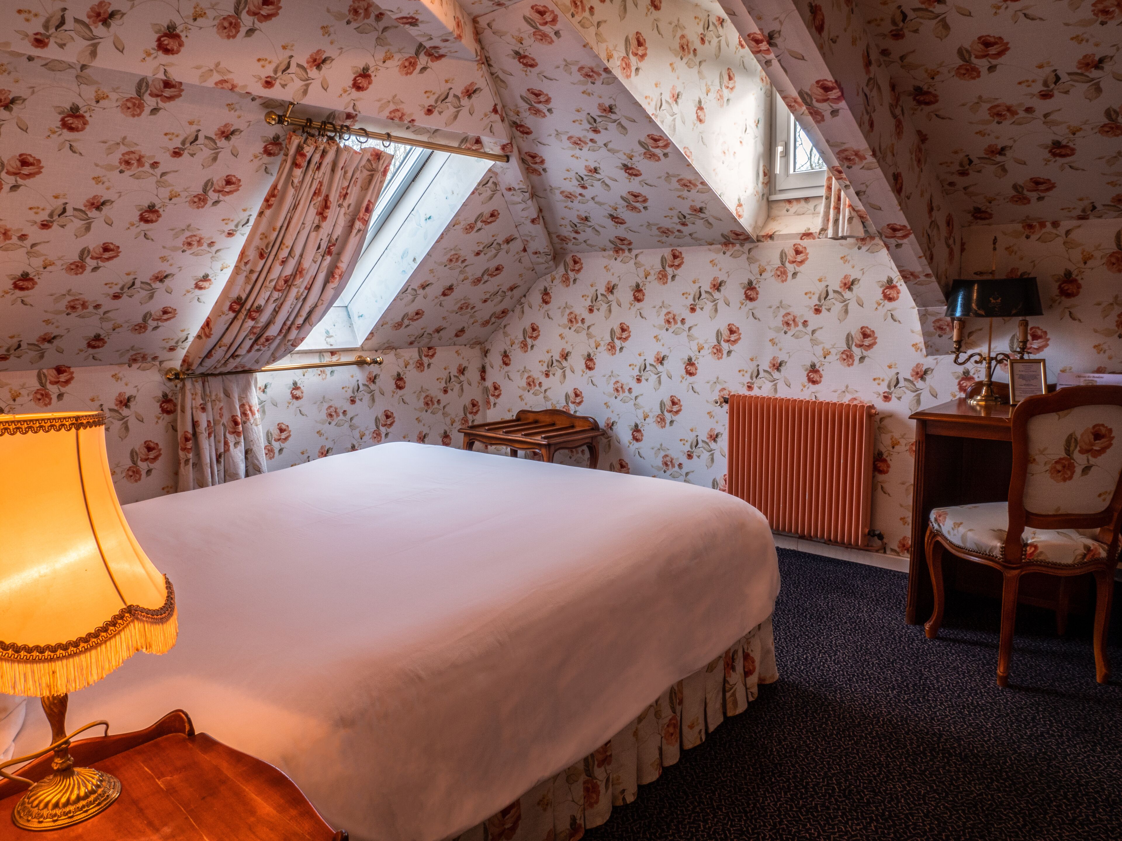 A bedroom with a large bed, a desk with a lamp, a chair, a radiator, and floral wallpaper.