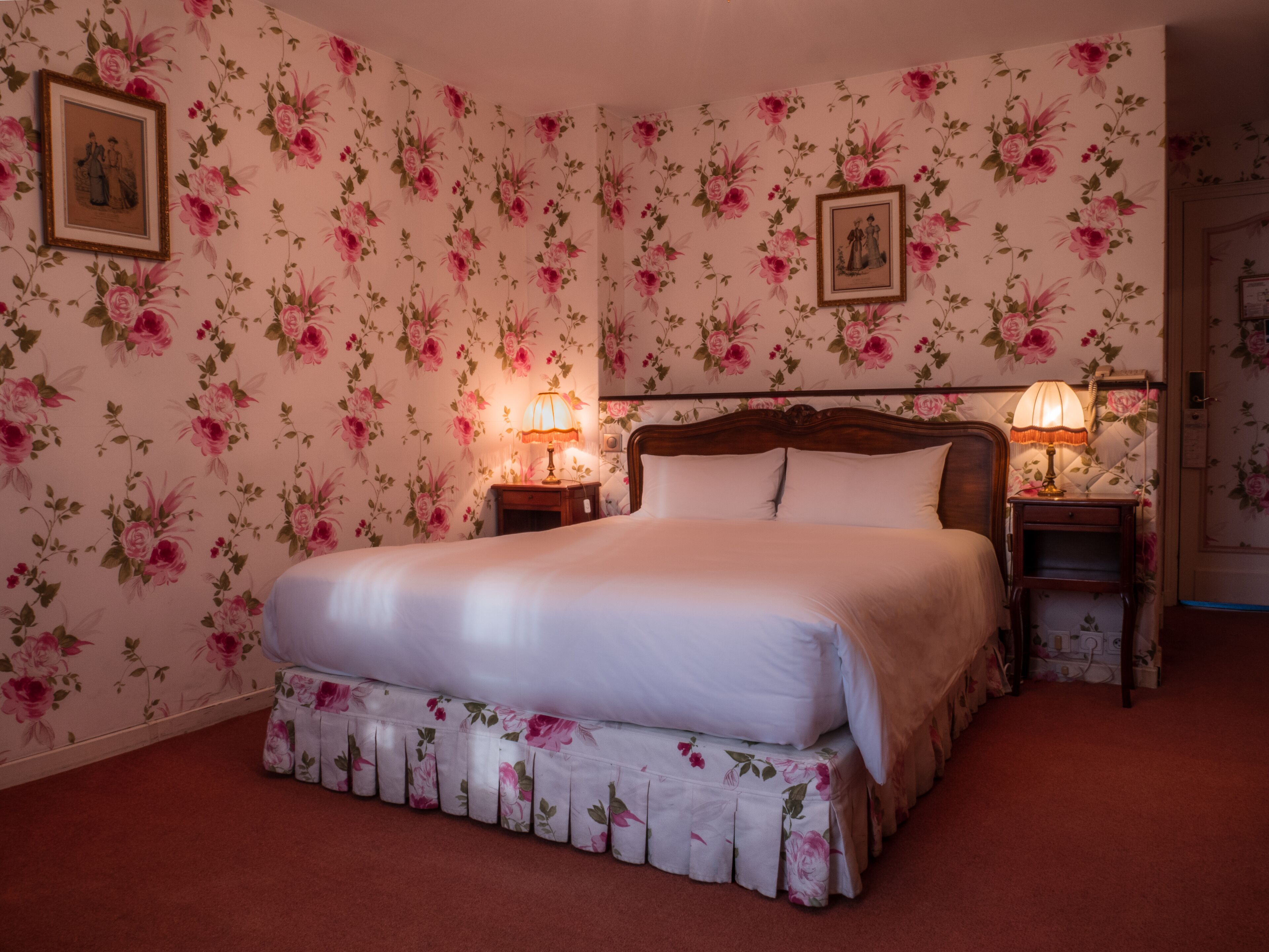 A bedroom with floral wallpaper, a large bed, two bedside tables with lamps, and framed pictures on the walls.