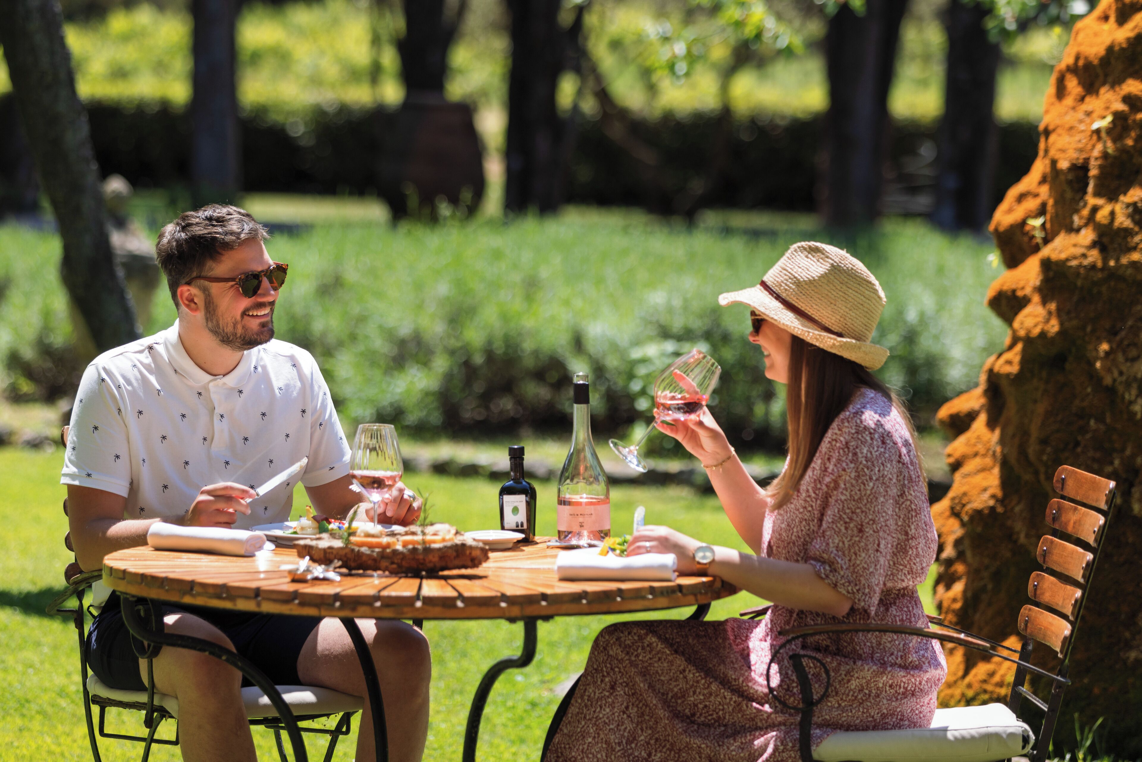 A couple dining at an outdoor table with a bottle of wine, a glass, and a plate of food.