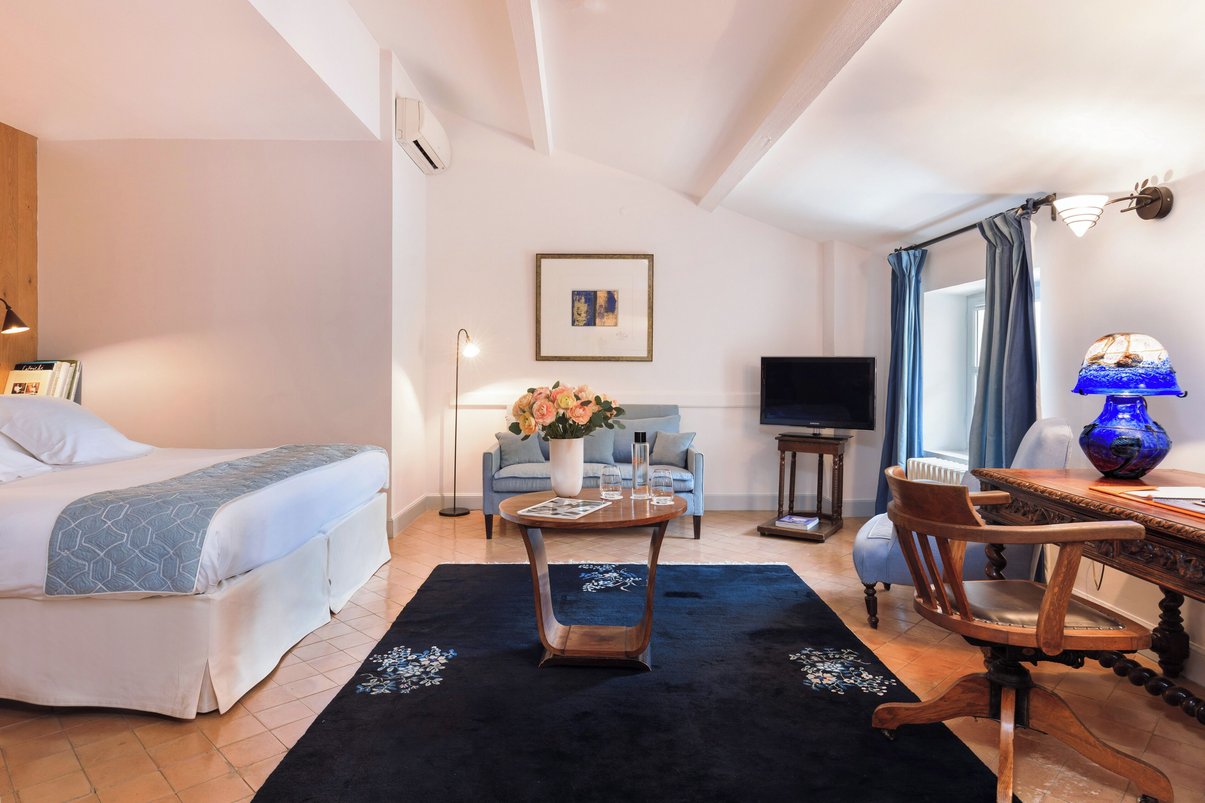 A hotel room with a bed, a sofa, a coffee table, a TV, and a vase with flowers.