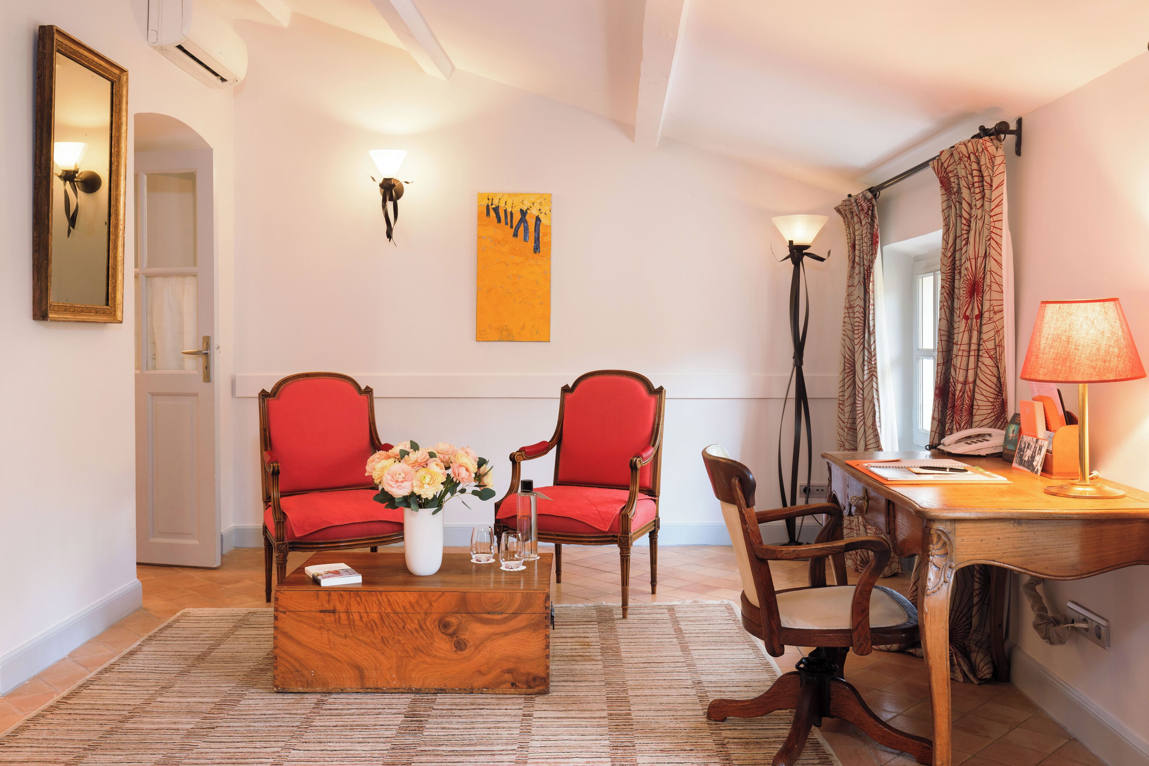 Una habitación con dos sillas rojas, una mesa de centro de madera, un escritorio con una lámpara, un florero con flores y un cuadro en la pared.