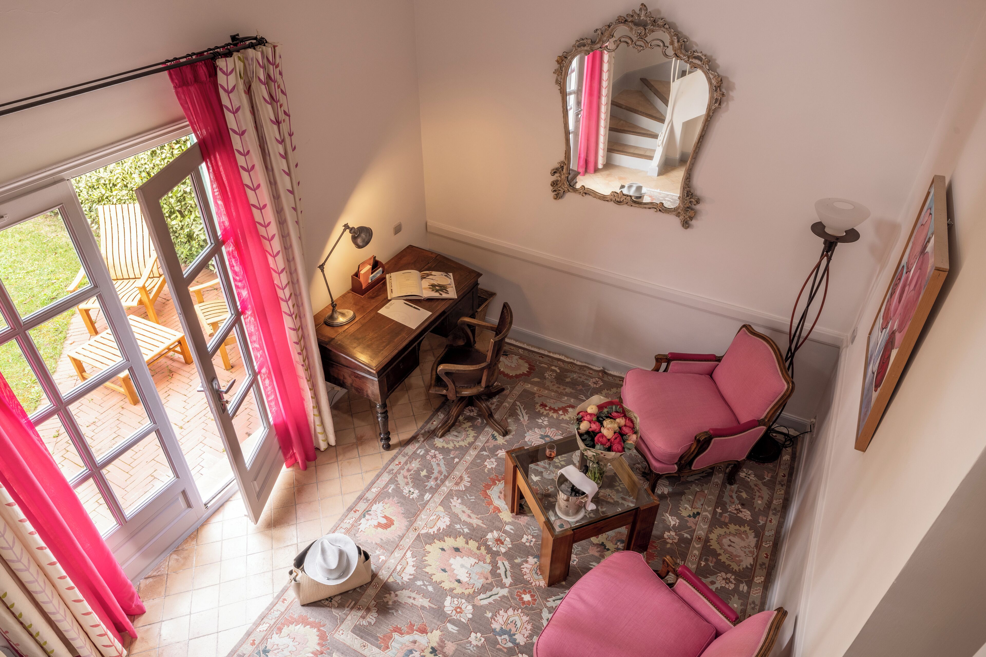Une pièce chaleureuse avec un bureau en bois, une chaise rose, un miroir et une vue sur l’extérieur grâce à une porte vitrée.
