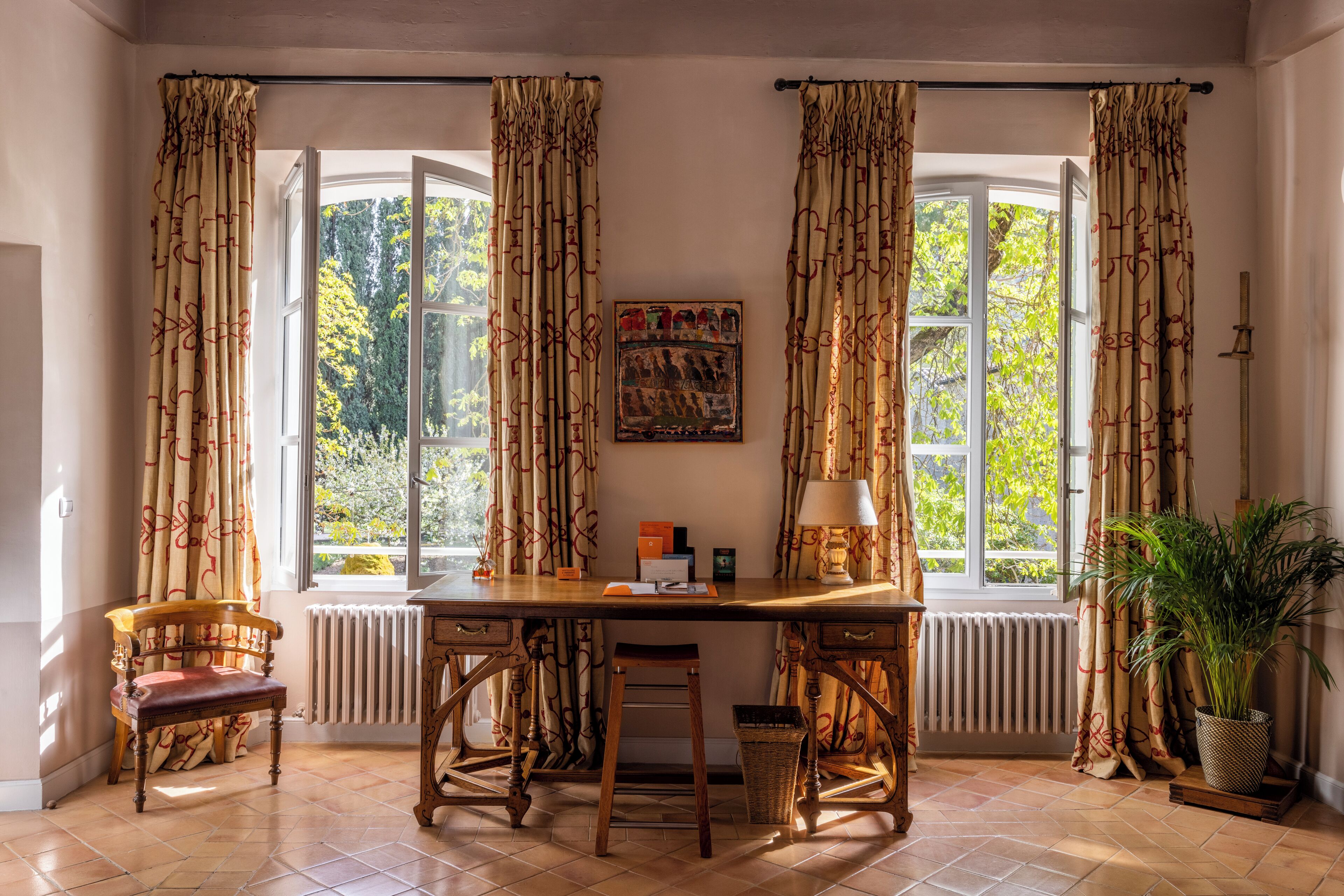 Una habitación con un escritorio de madera, dos sillas, un radiador, una ventana con cortinas, una planta en maceta y un cuadro en la pared.