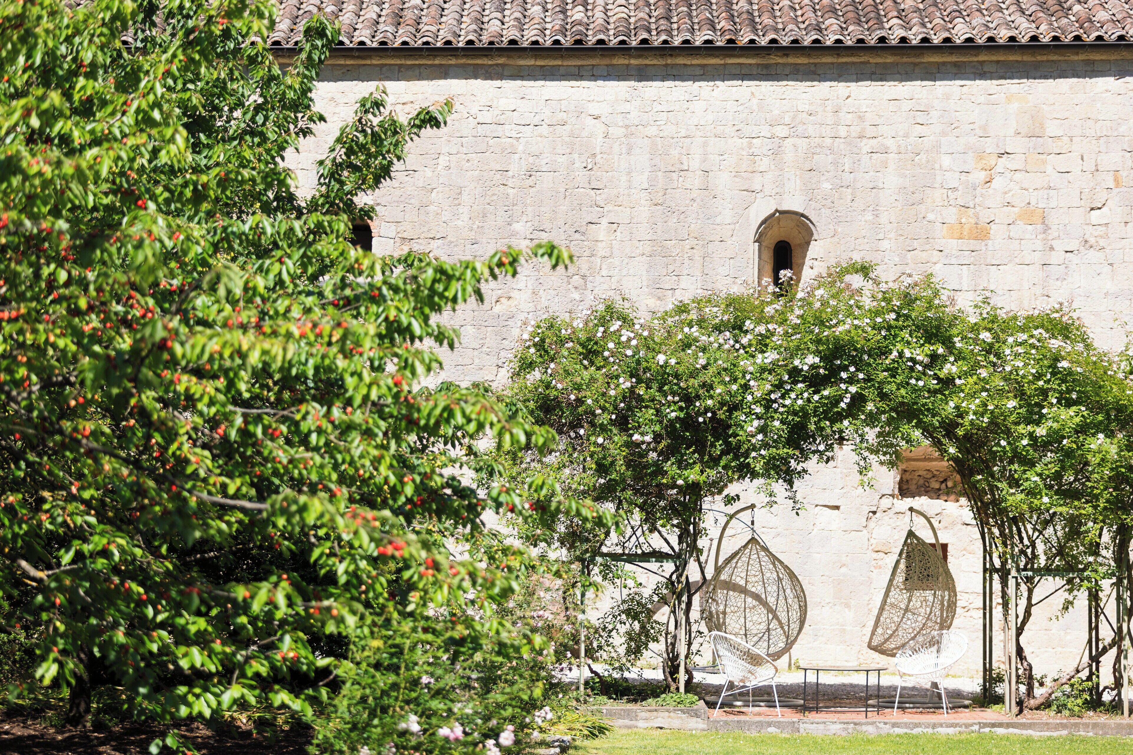 A stone building with a tiled roof, surrounded by greenery and two white chairs under a pergola.