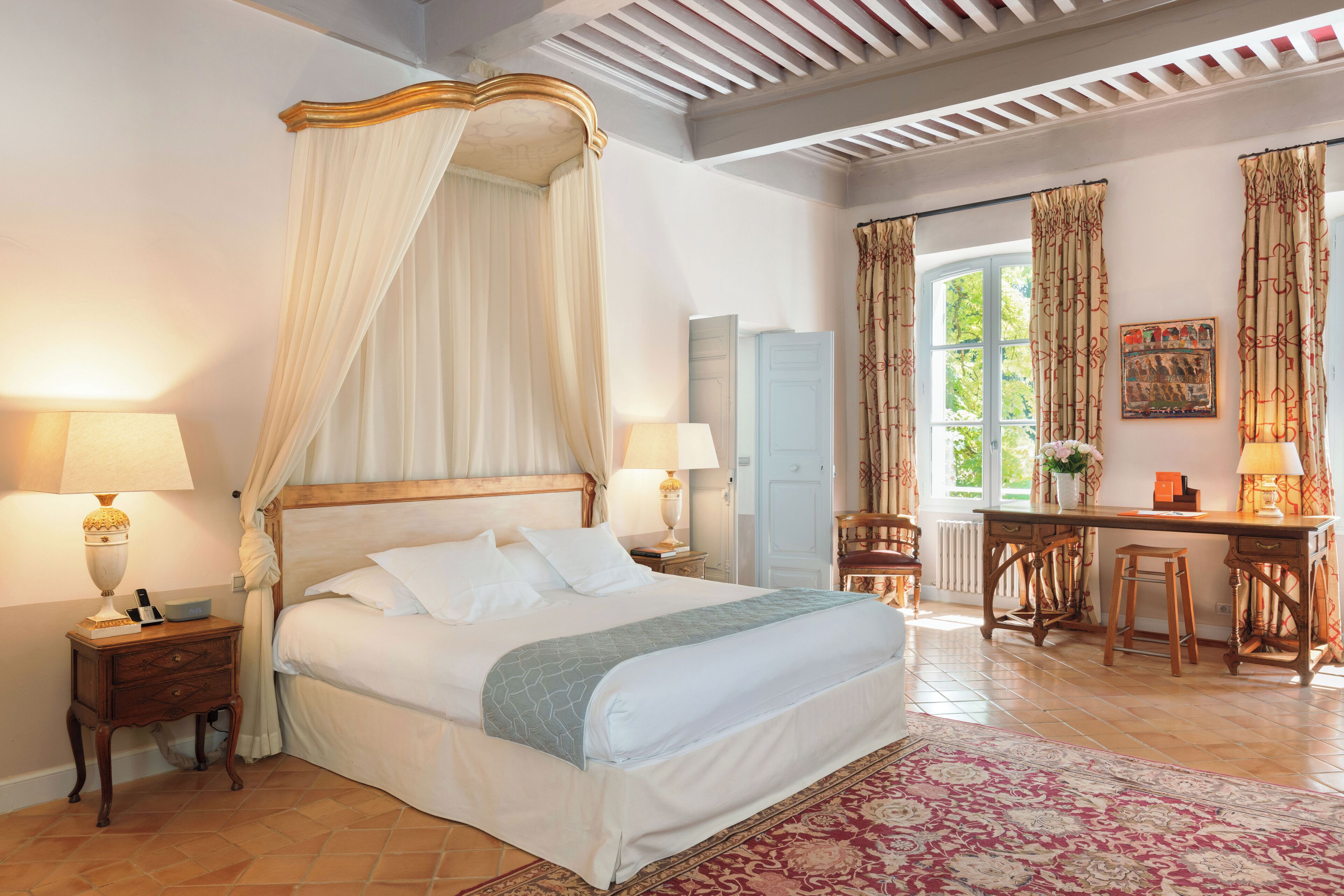 A bedroom with a four-poster bed, a desk with two chairs, and a view of the outdoors through a window.