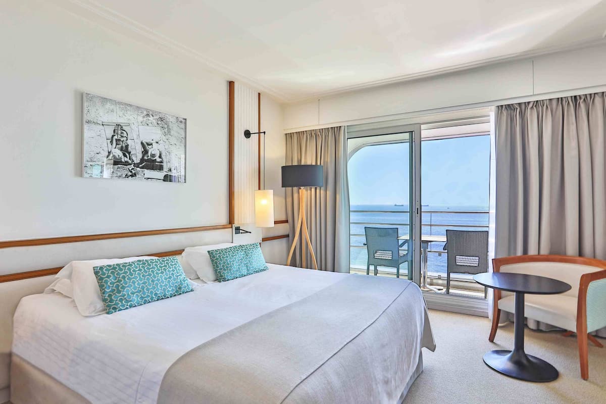 A hotel room with a large bed, a small table, a chair, a balcony with a view of the sea, and a painting on the wall.