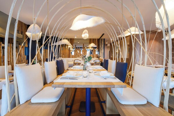 Un restaurante con una larga mesa de madera, bancos blancos acolchados y lámparas blancas colgantes.