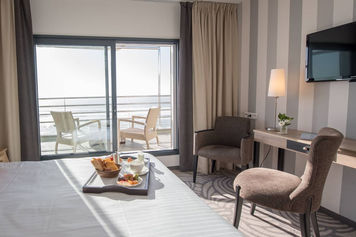 A hotel room with a bed, a desk, two chairs, a TV, and a view of the ocean.