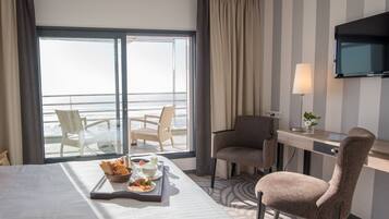 A hotel room with a bed, a desk, two chairs, a TV, and a view of the ocean.