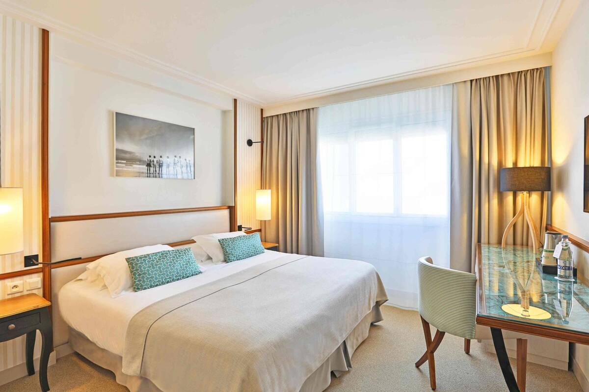 A hotel room with a large bed, a desk with a lamp, and a window with curtains.