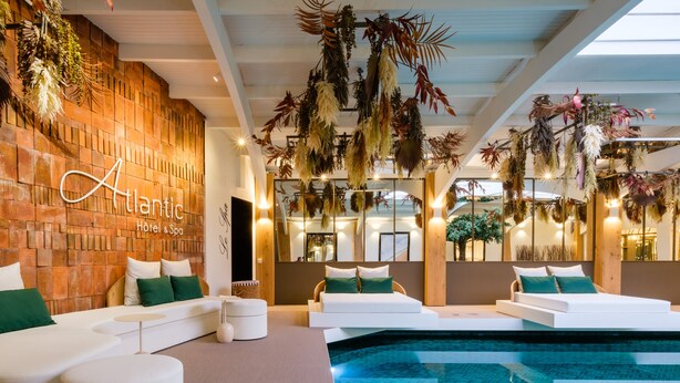 Un interior moderno de hotel con piscina, sofás blancos y plantas colgantes.