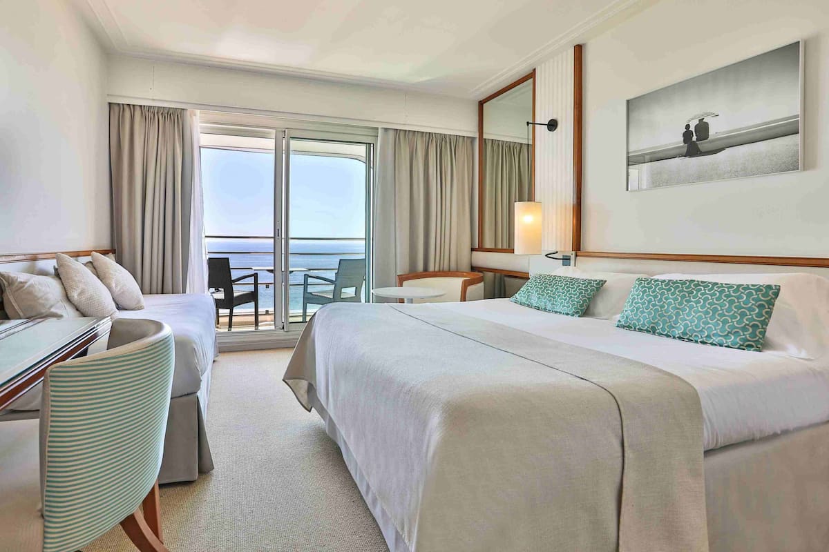 A hotel room with a bed, a desk, a chair, a small table, and a balcony with a view of the sea.