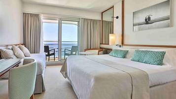 Quarto de hotel com cama, escrivaninha, cadeira, mesa pequena e varanda com vista para o mar.