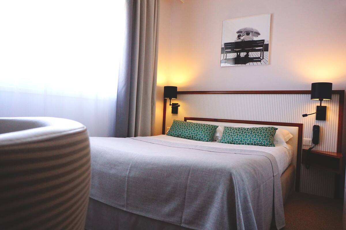 A neatly made bed with a grey bedspread and two patterned pillows, flanked by two bedside tables with lamps, in a room with a large window and a framed artwork on the wall.