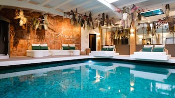 Un área de piscina interior moderna con una piscina azul cristalina, sofás blancos con cojines verdes y una pared de madera con plantas colgantes.