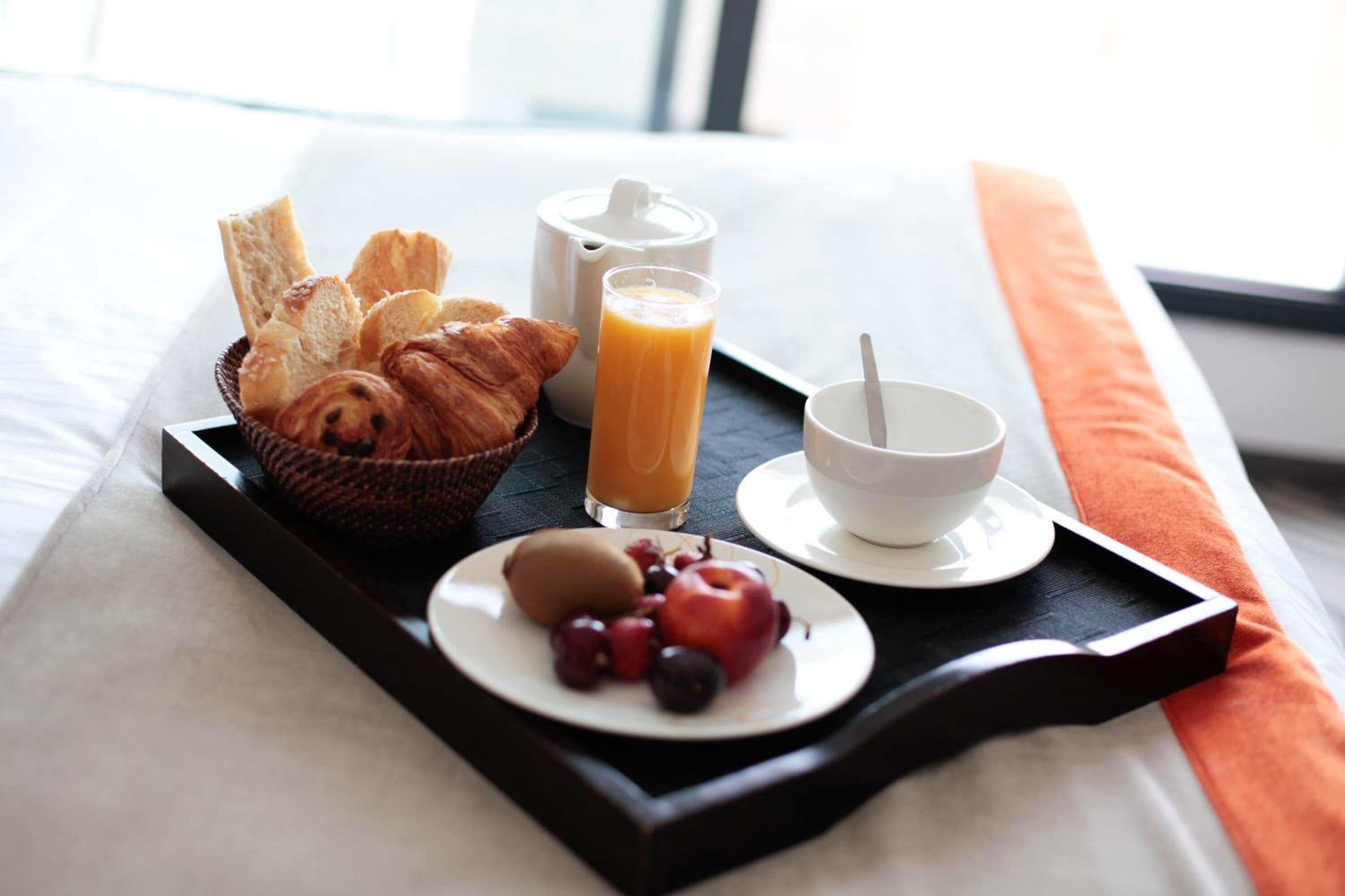 A breakfast tray with croissants, a jug of orange juice, a cup of coffee, and a plate of fruit on a bed.