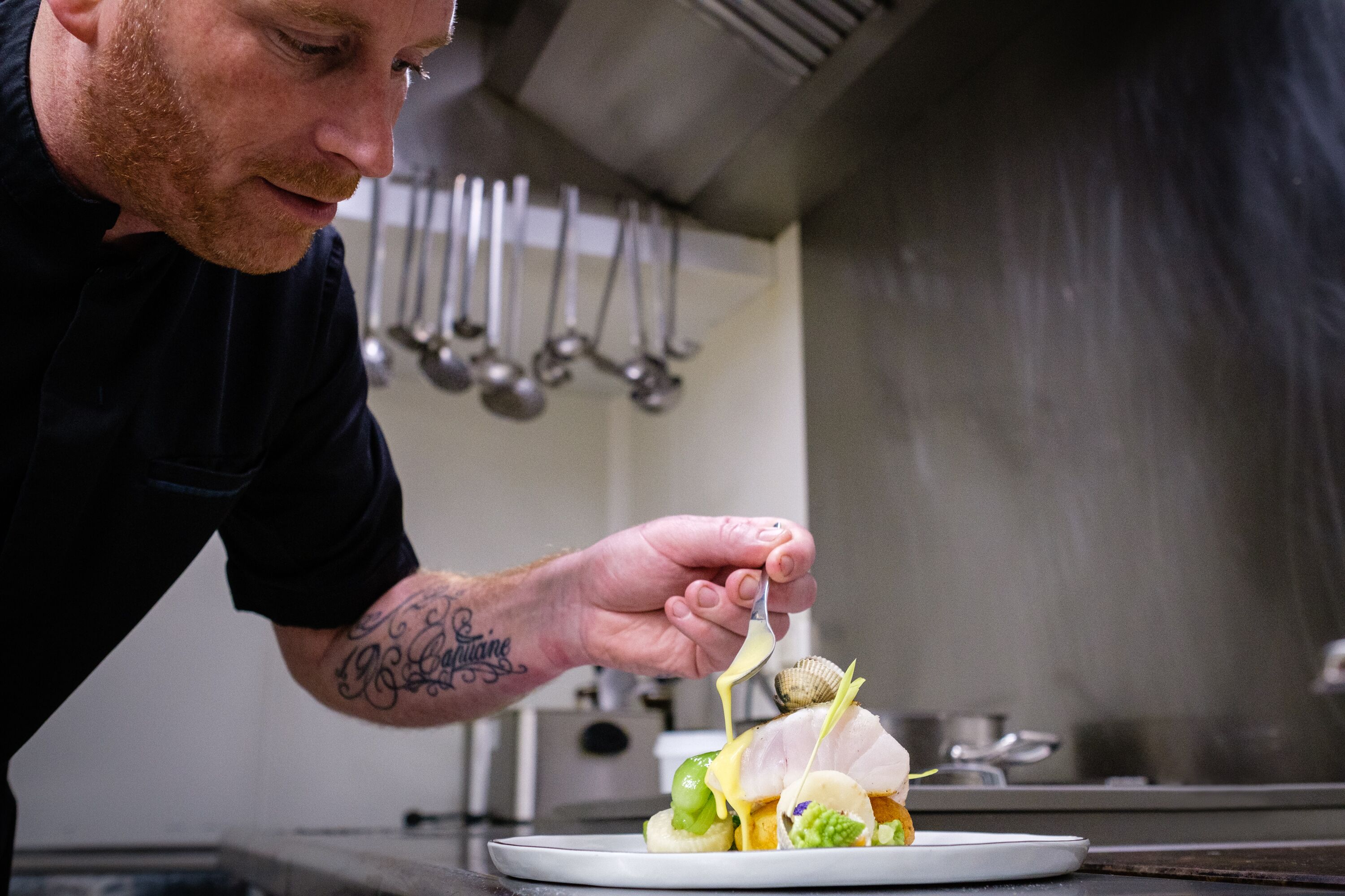 Um chef preparando um prato na cozinha, com utensílios pendurados.