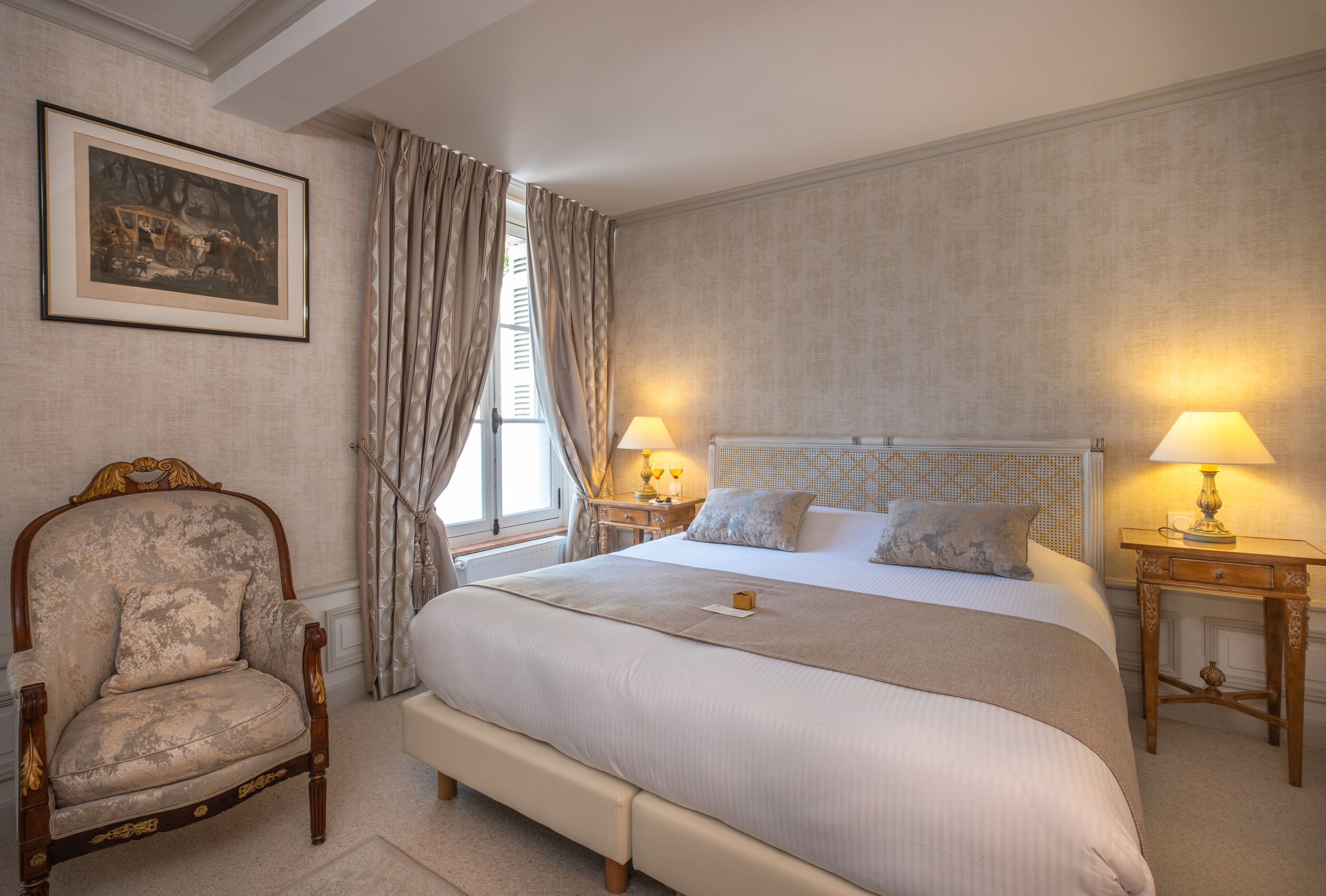 A bedroom with a large bed, two bedside tables with lamps, a chair, and a framed picture on the wall.