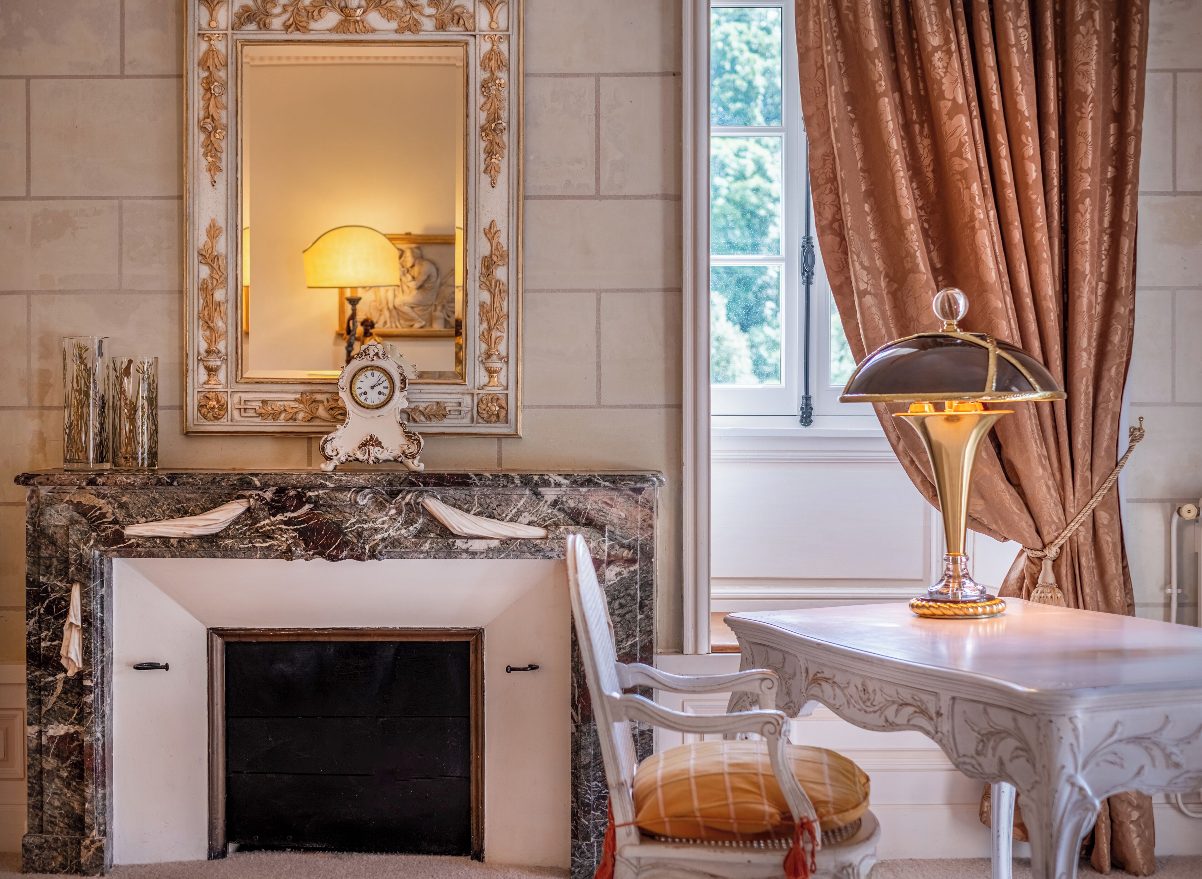 A room with a marble fireplace, a white ornate table, a white chair, a large mirror, and a window with curtains.