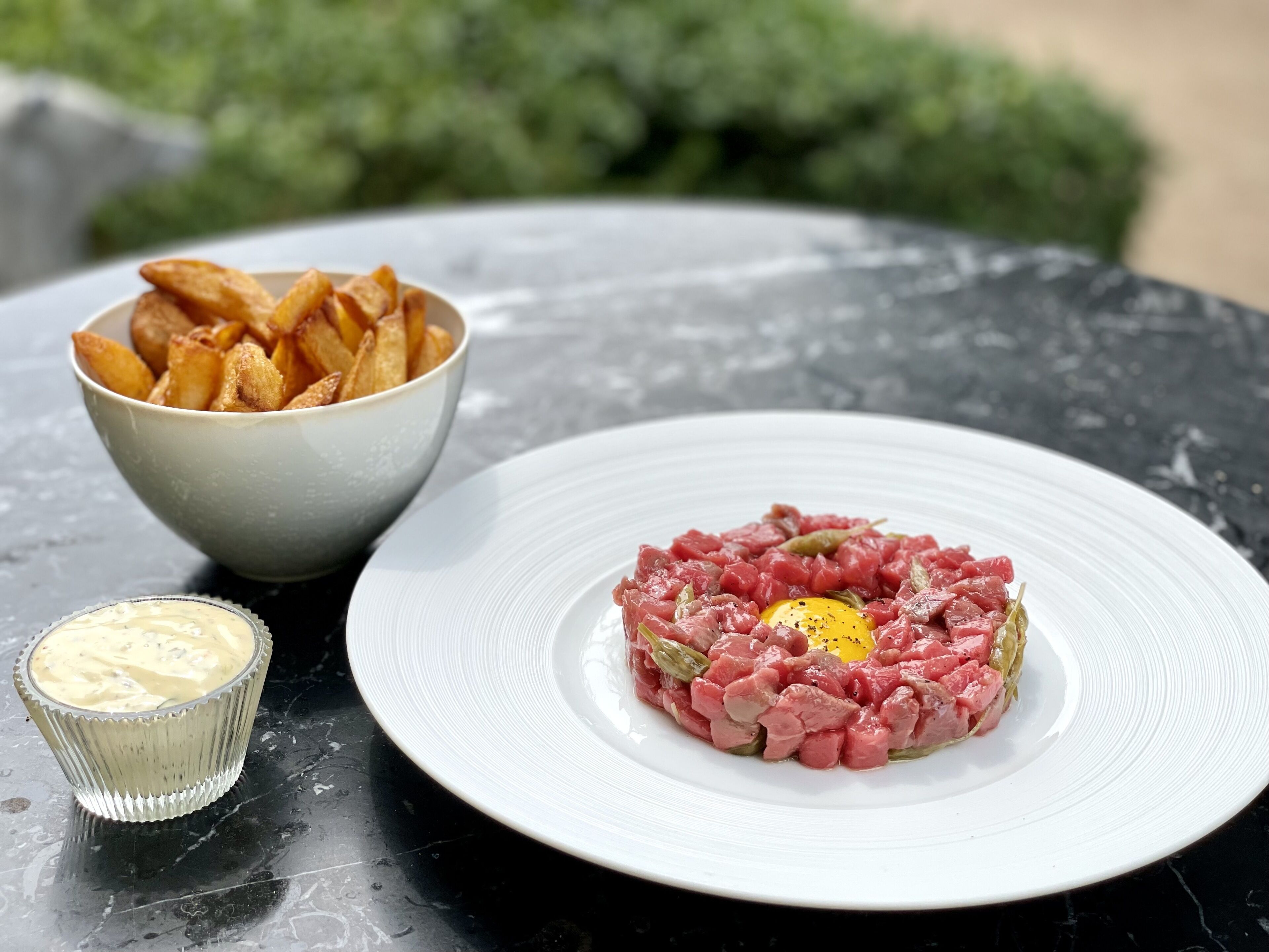 A plate of steak tartare with a raw egg yolk in the center, served with a side of potato chips and a small bowl of sauce.