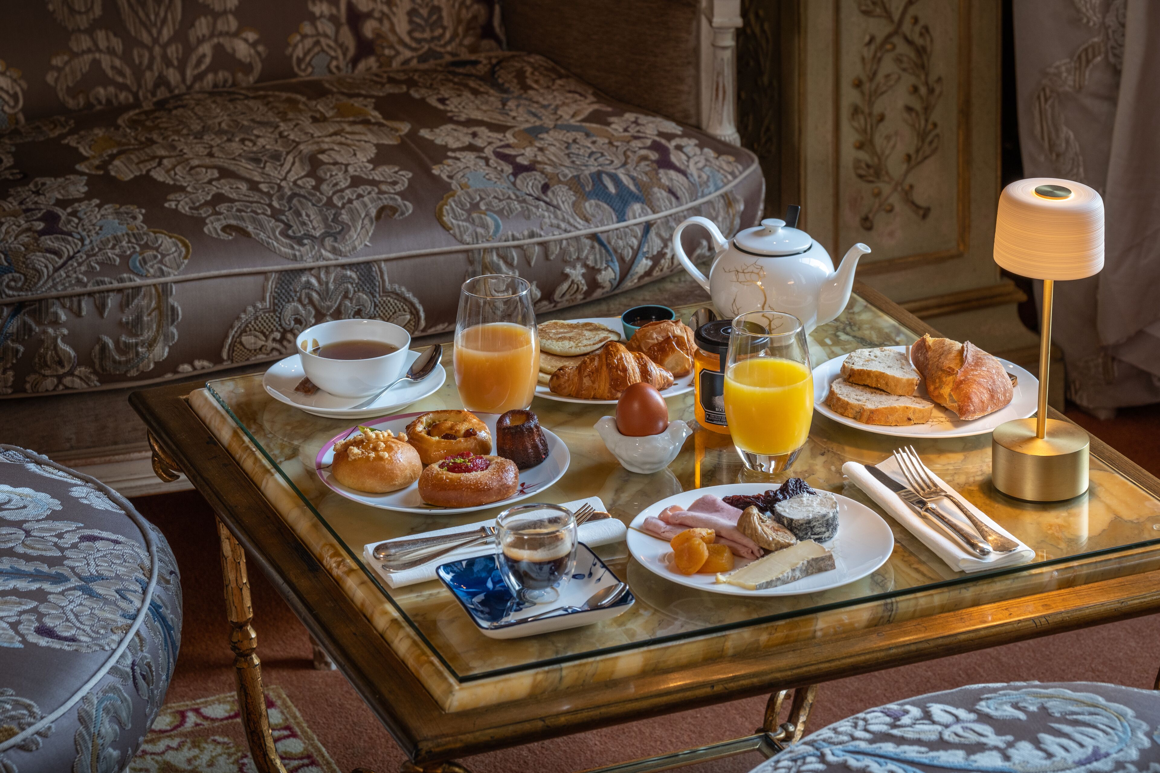 A breakfast table with various food items, a teapot, and a lamp.