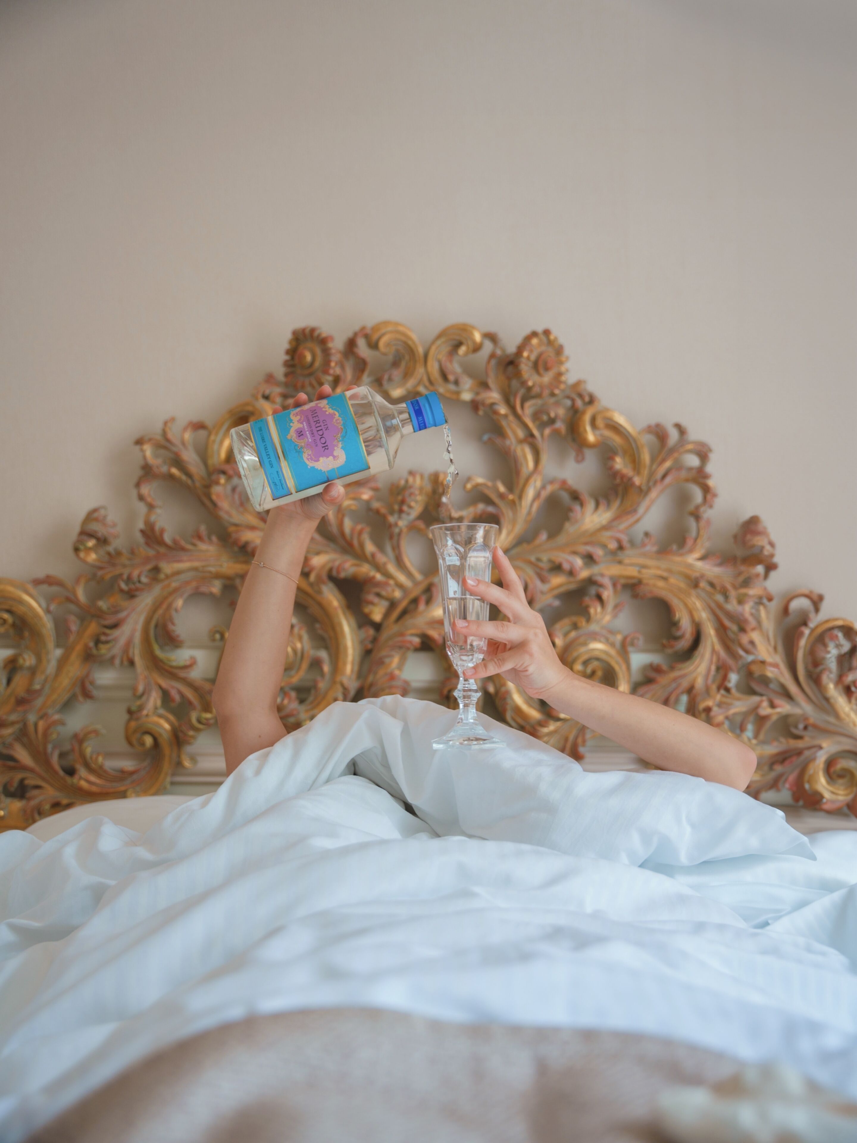 Una persona vertiendo un líquido transparente de una botella en un vaso sobre una cama con cabecera decorada.