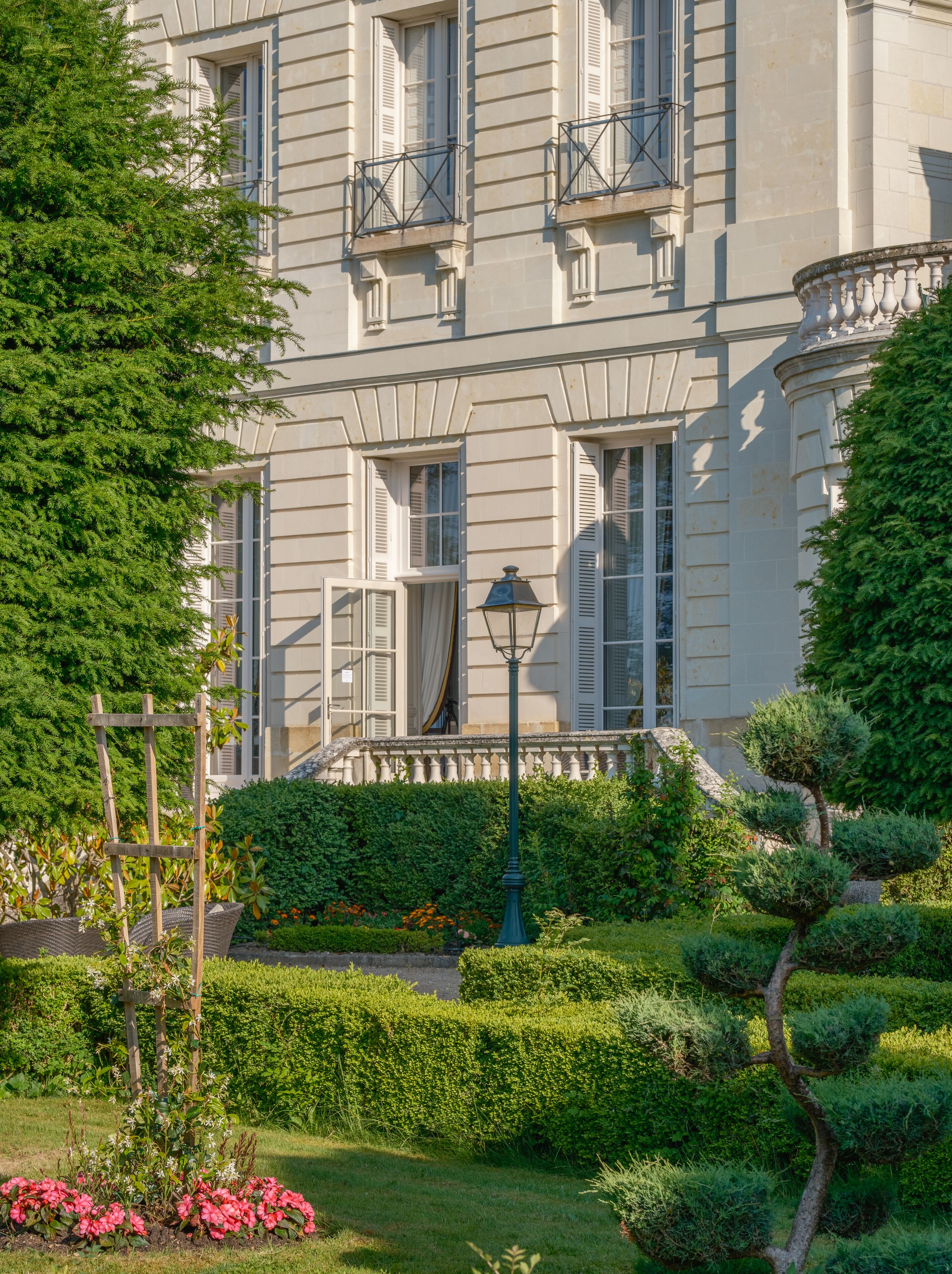 Un immeuble à plusieurs étages doté de balcons, un jardin avec des haies et des fleurs, et un lampadaire.