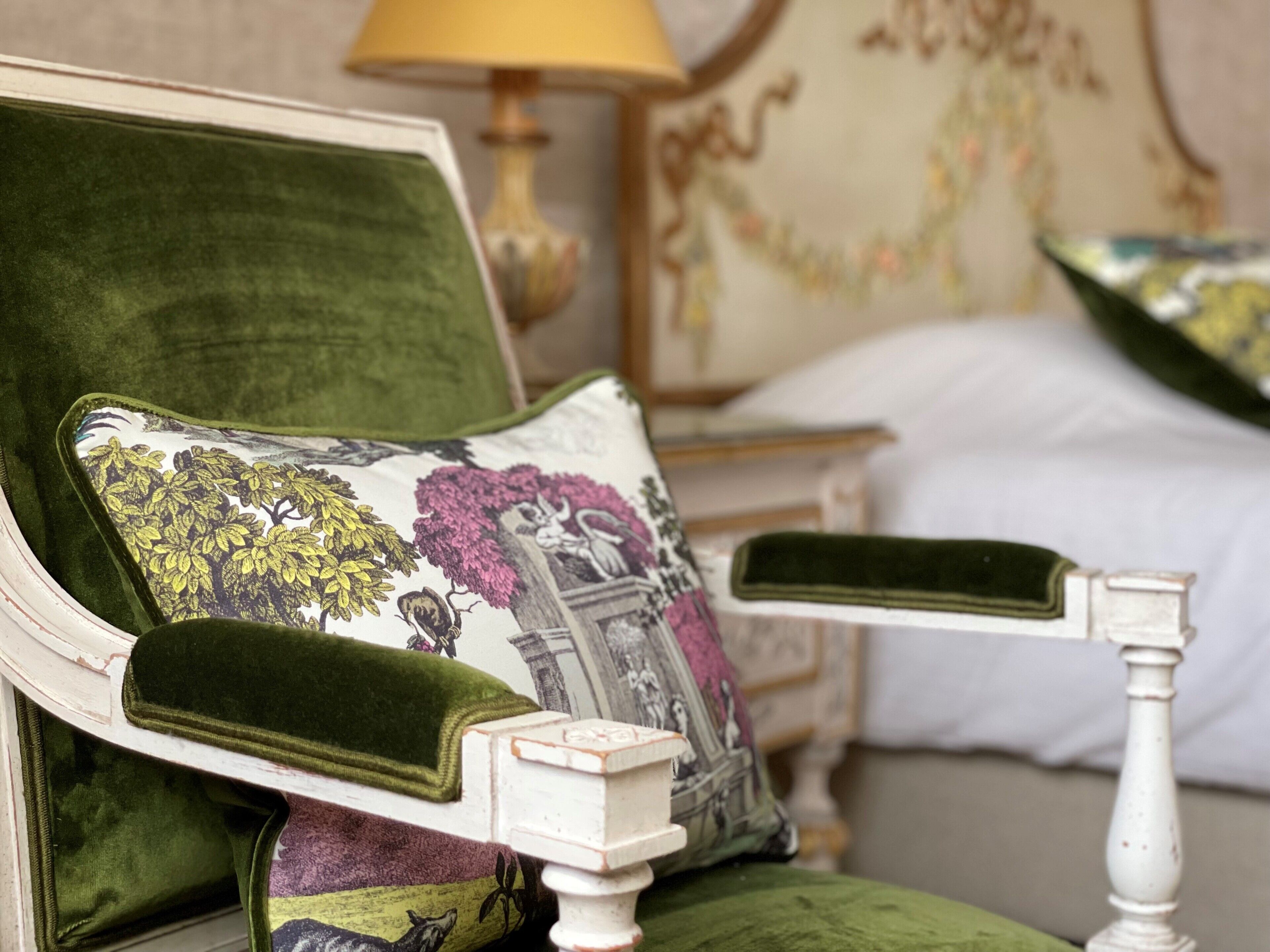 A bed with a green upholstered headboard, a decorative pillow with floral and animal prints, and a white bedside table with a lamp.