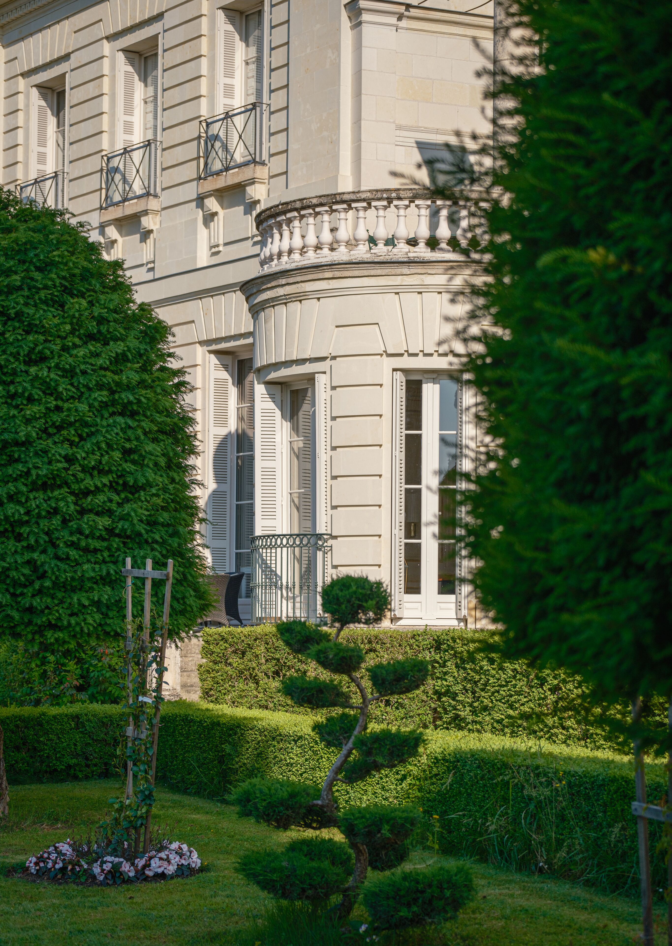 Un edificio clásico con arquitectura simétrica, un jardín bien cuidado con setos y un pequeño árbol, y un balcón con barandillas metálicas.