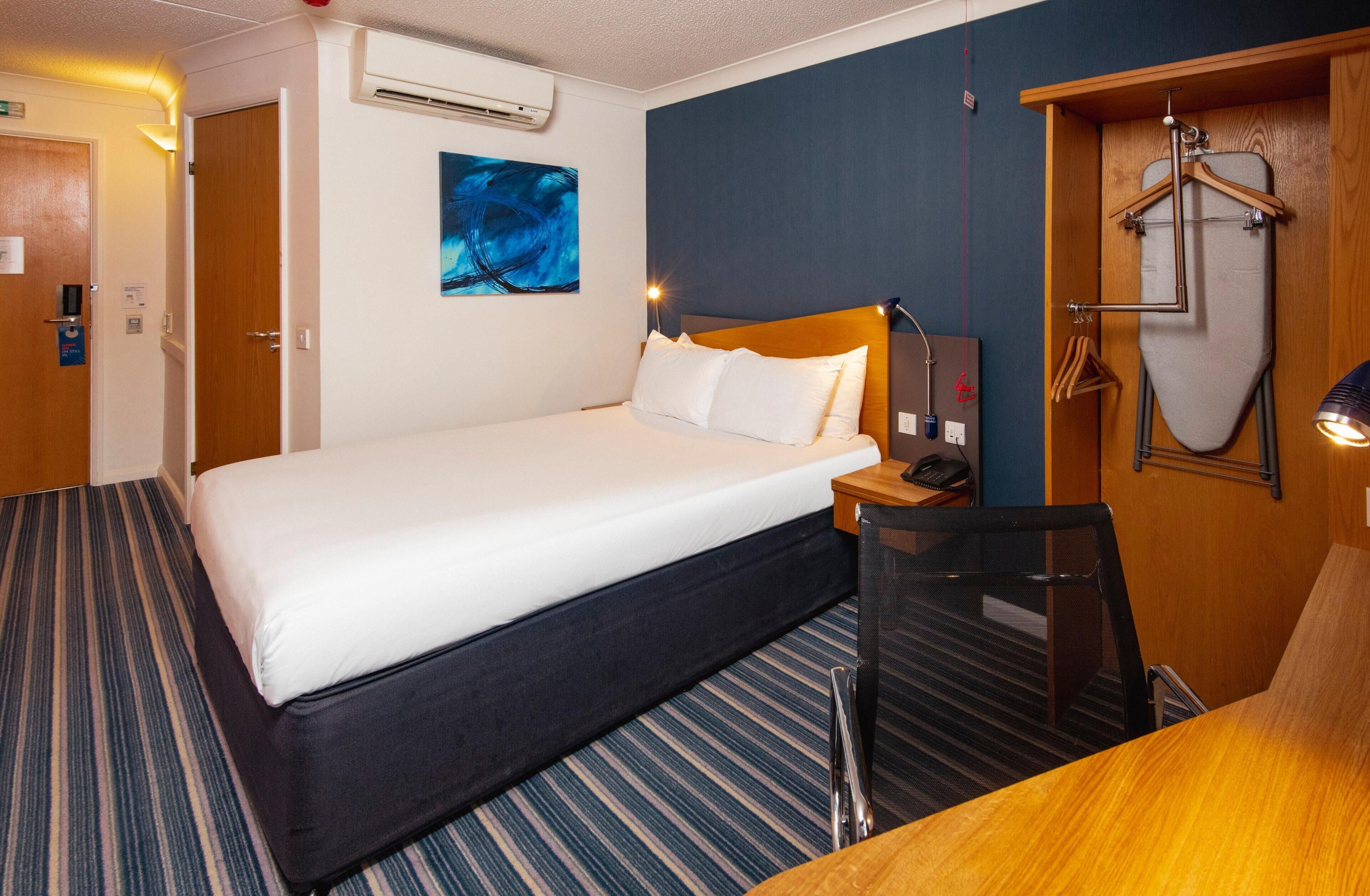A hotel room with a bed, a desk, a chair, an ironing board, and a painting on the wall.