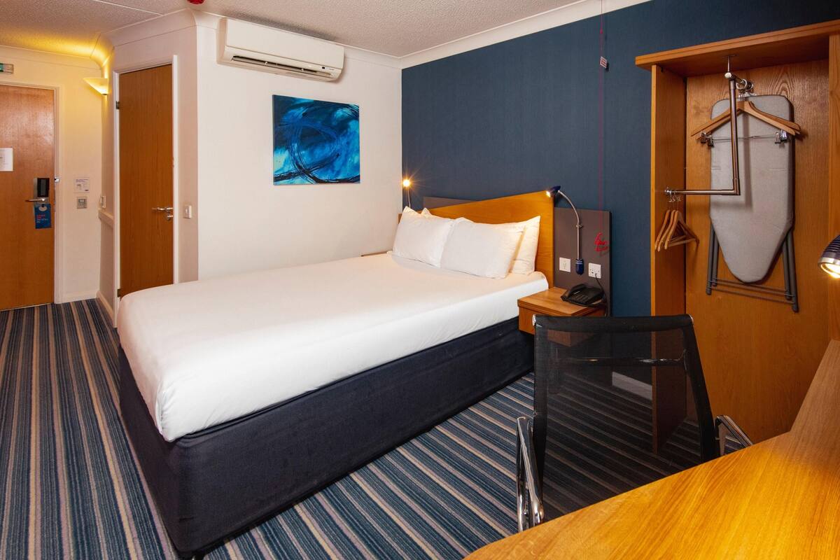 A hotel room with a large bed, a desk, an ironing board, and a painting on the wall.